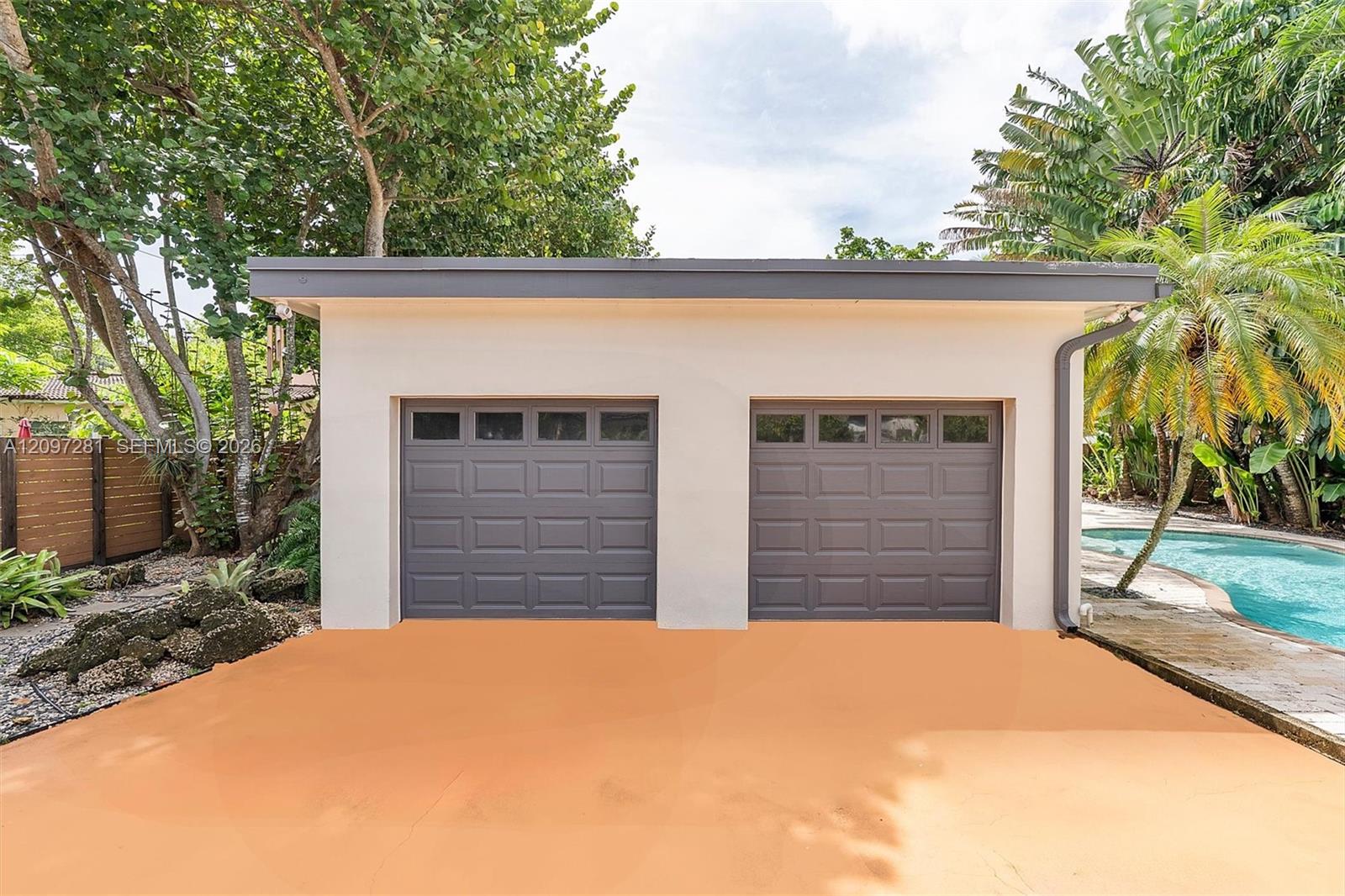 House exterior with two gray garage doors, a peach-colored driveway, and a pool beside tropical palms.