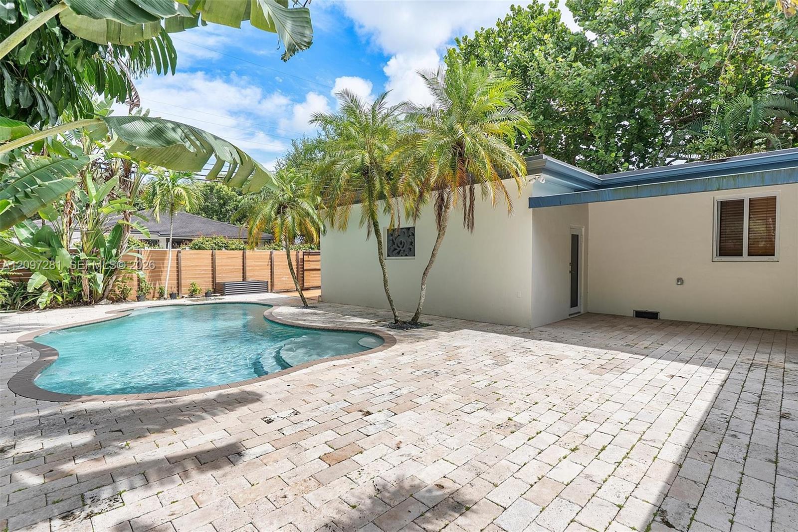 Curved backyard pool beside a light-colored house, surrounded by palms and a wide paved patio.