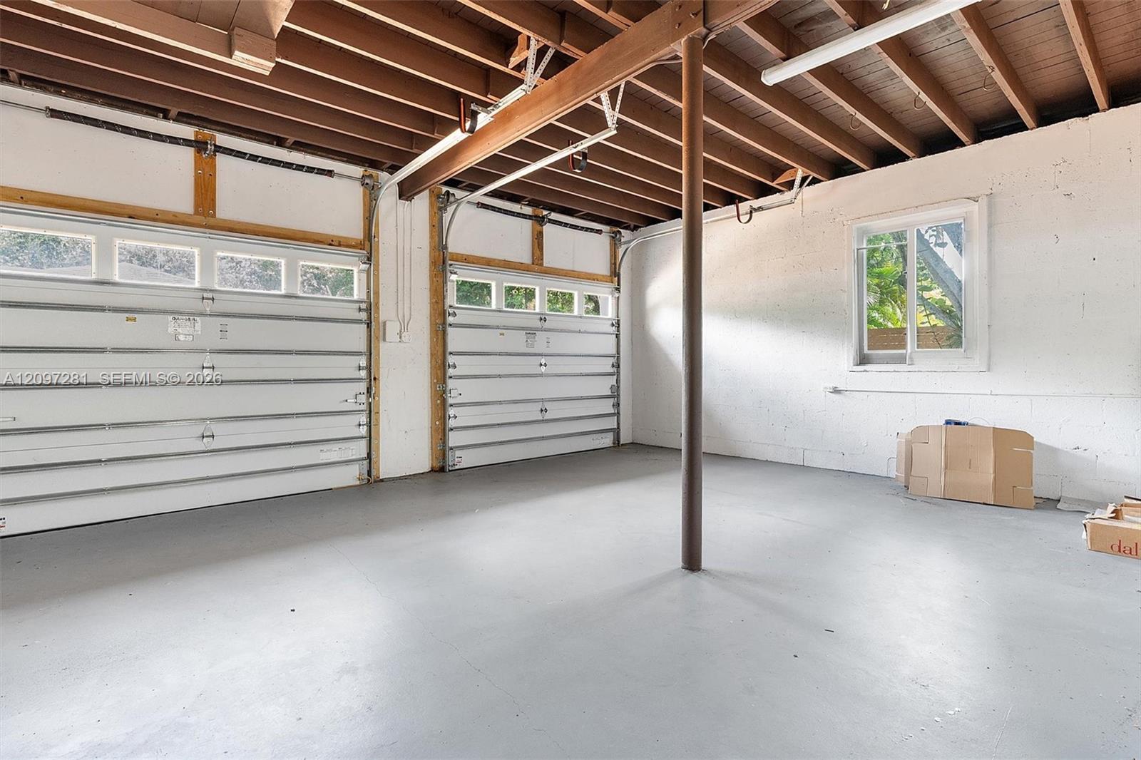 Mostly empty two-car garage with exposed wooden beams, a concrete floor, and boxes by the wall.