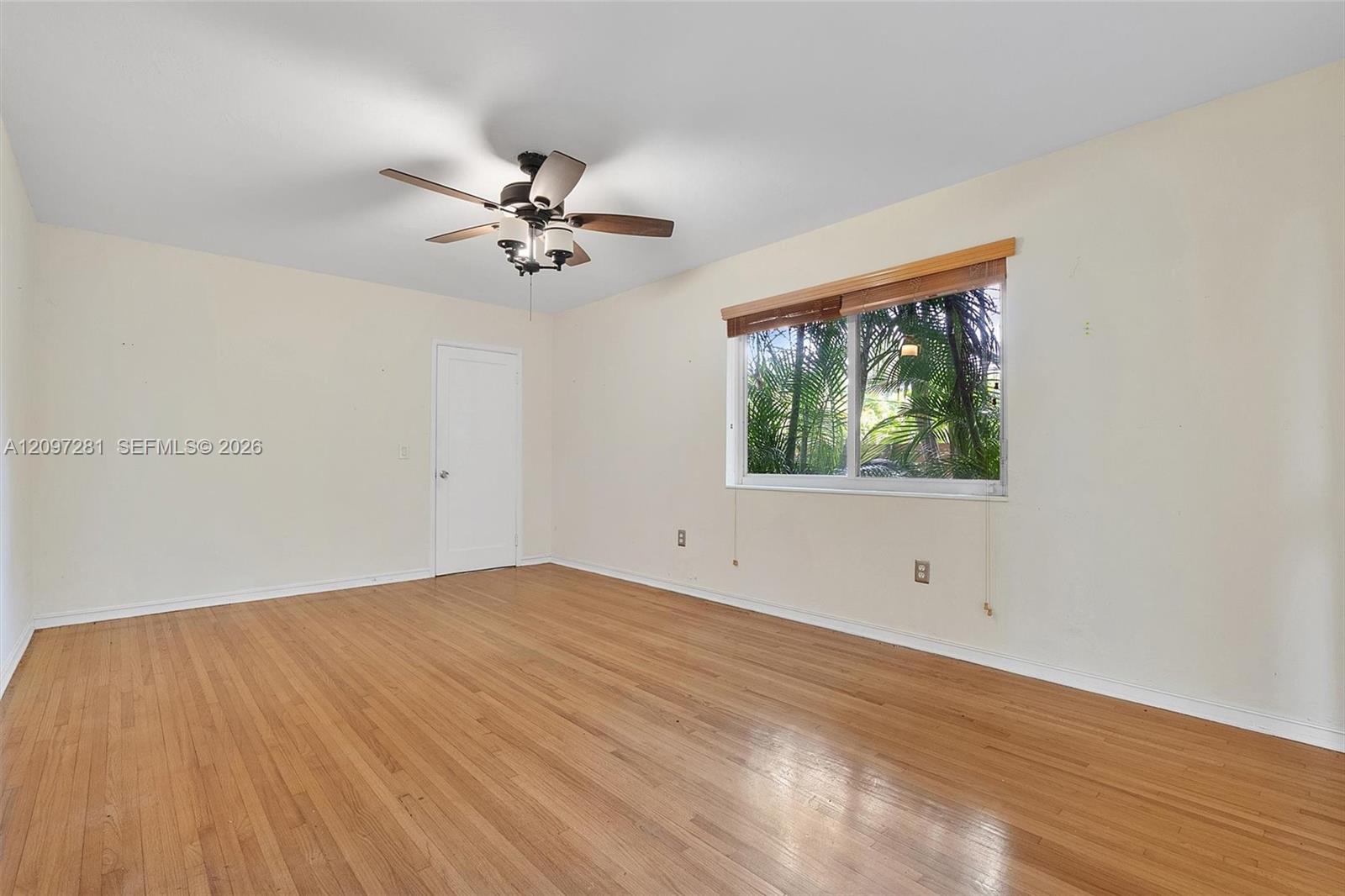 Empty room with hardwood floors, a ceiling fan, and a window overlooking palm foliage.