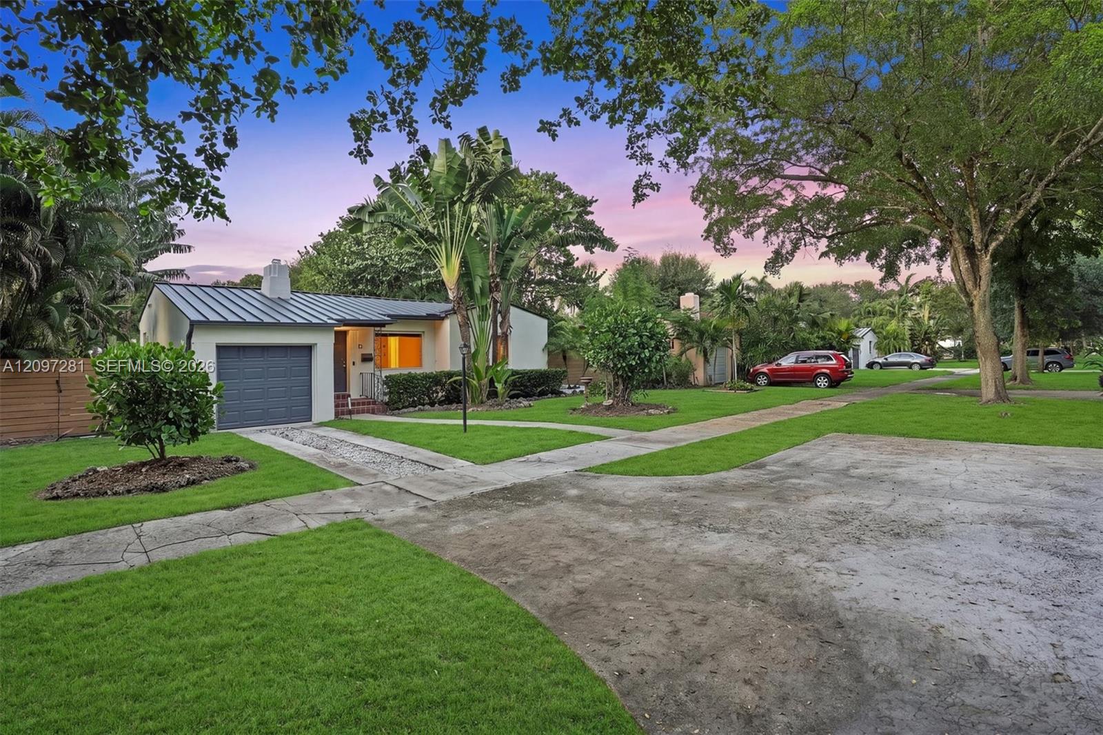 Single-story home with a dark metal roof, attached garage, and tropical landscaping at dusk.