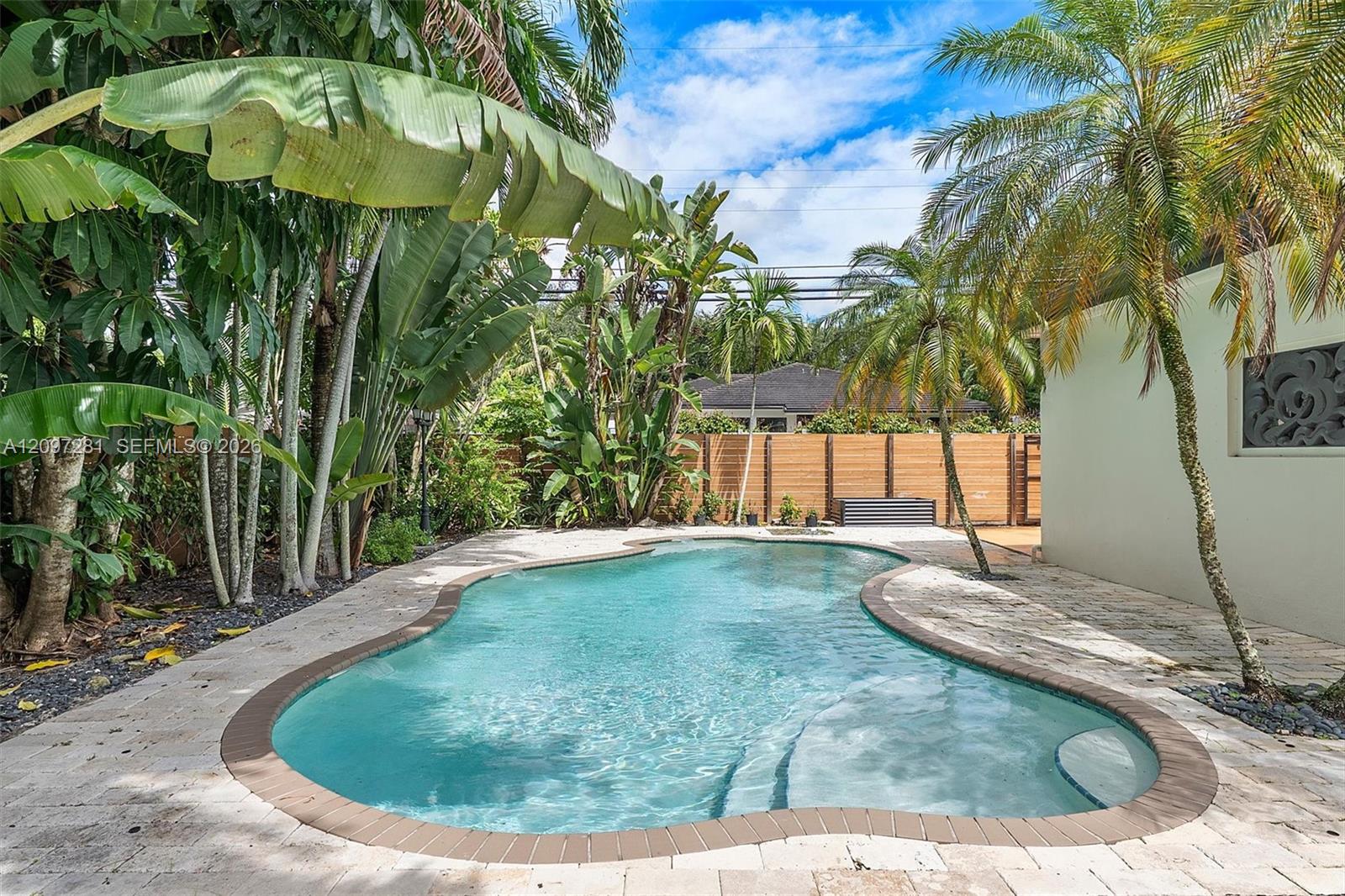 Curved backyard pool surrounded by tropical palms, stone paving, and a wooden privacy fence.