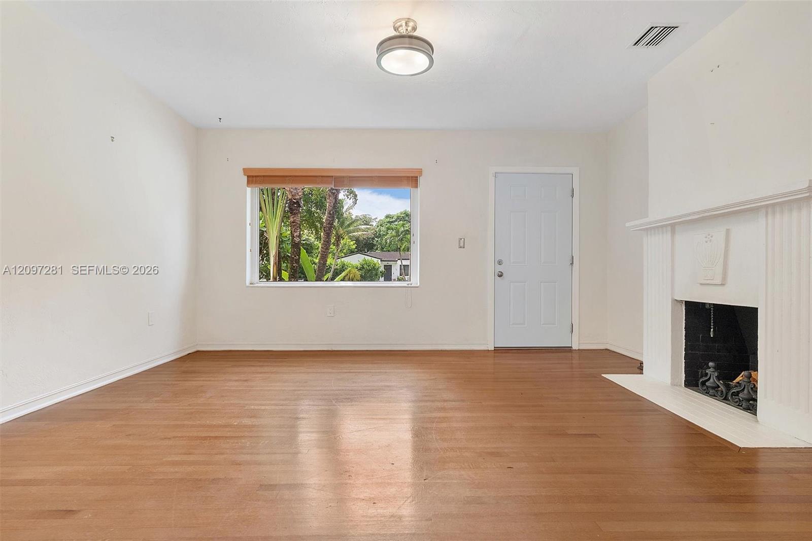 Vacant living room with wood floors, a white fireplace, and a window overlooking tropical greenery.