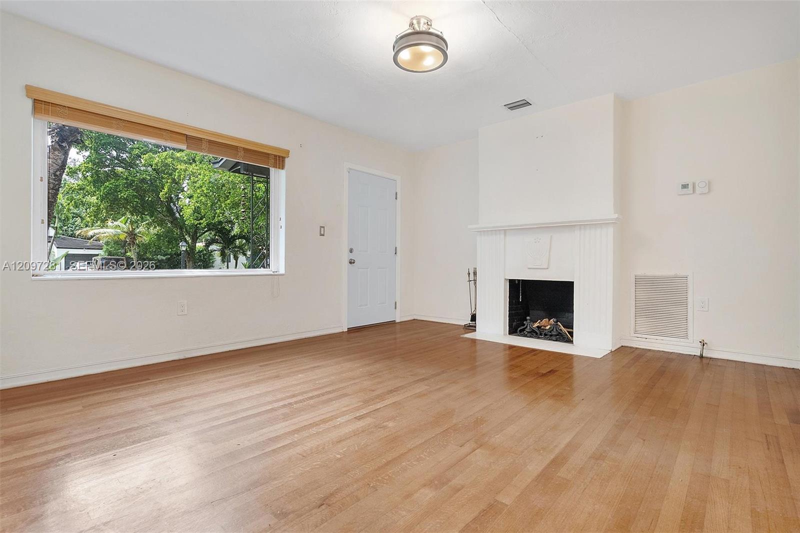 Empty living room with hardwood floors, a white fireplace, and a large window overlooking trees.