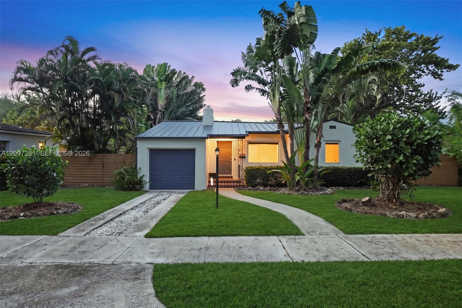 Single-story home with a blue garage door, metal roof, and tropical landscaping at dusk.