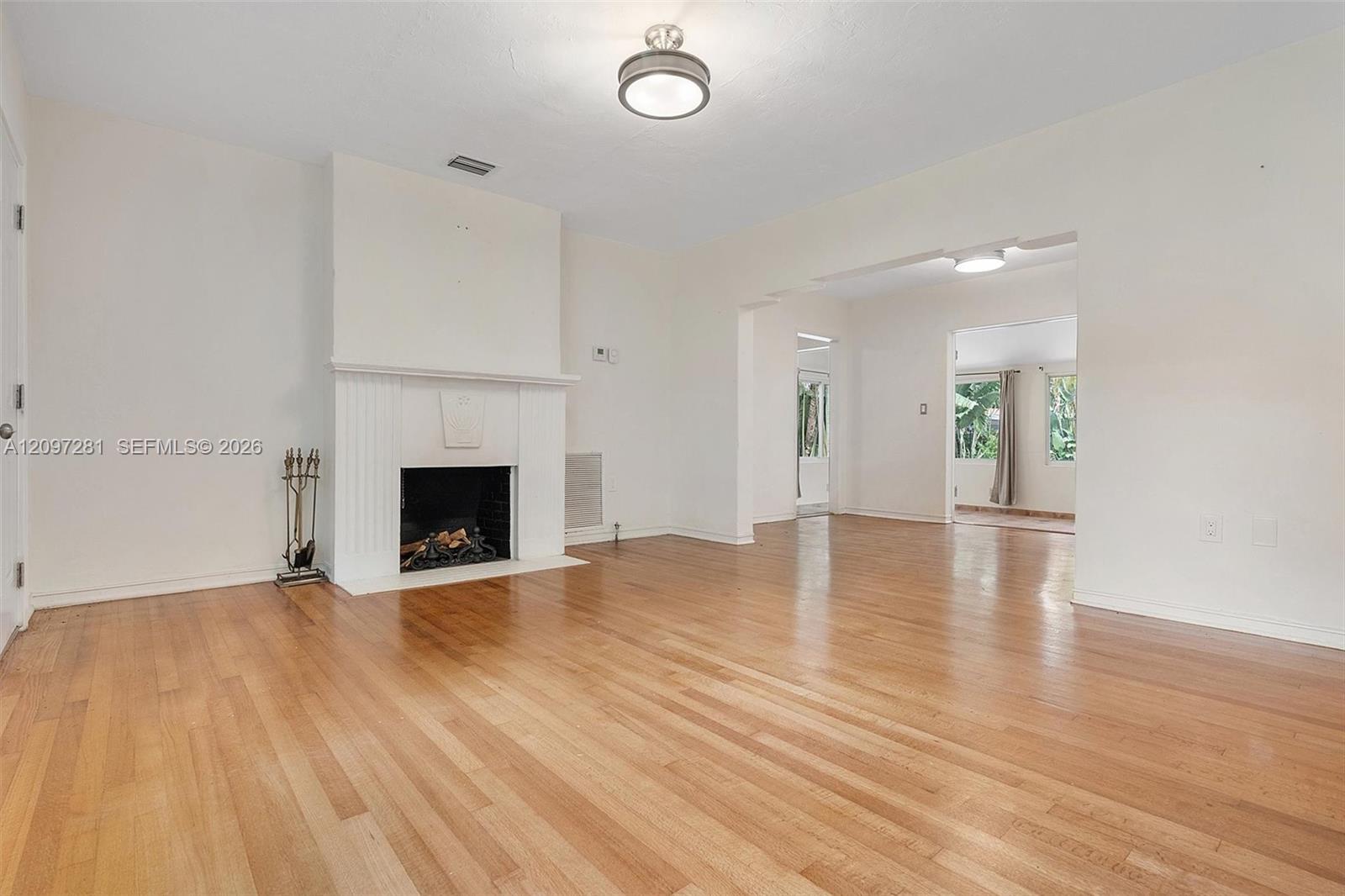 Vacant living room with polished hardwood floors, a white fireplace, and an open passage to a bright adjoining room.