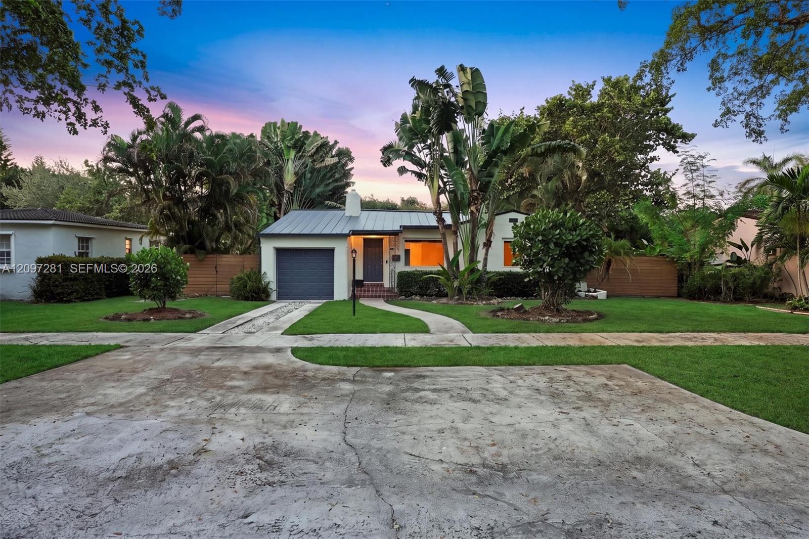 Single-story home with a dark metal roof, tropical landscaping, and warmly lit windows at dusk.