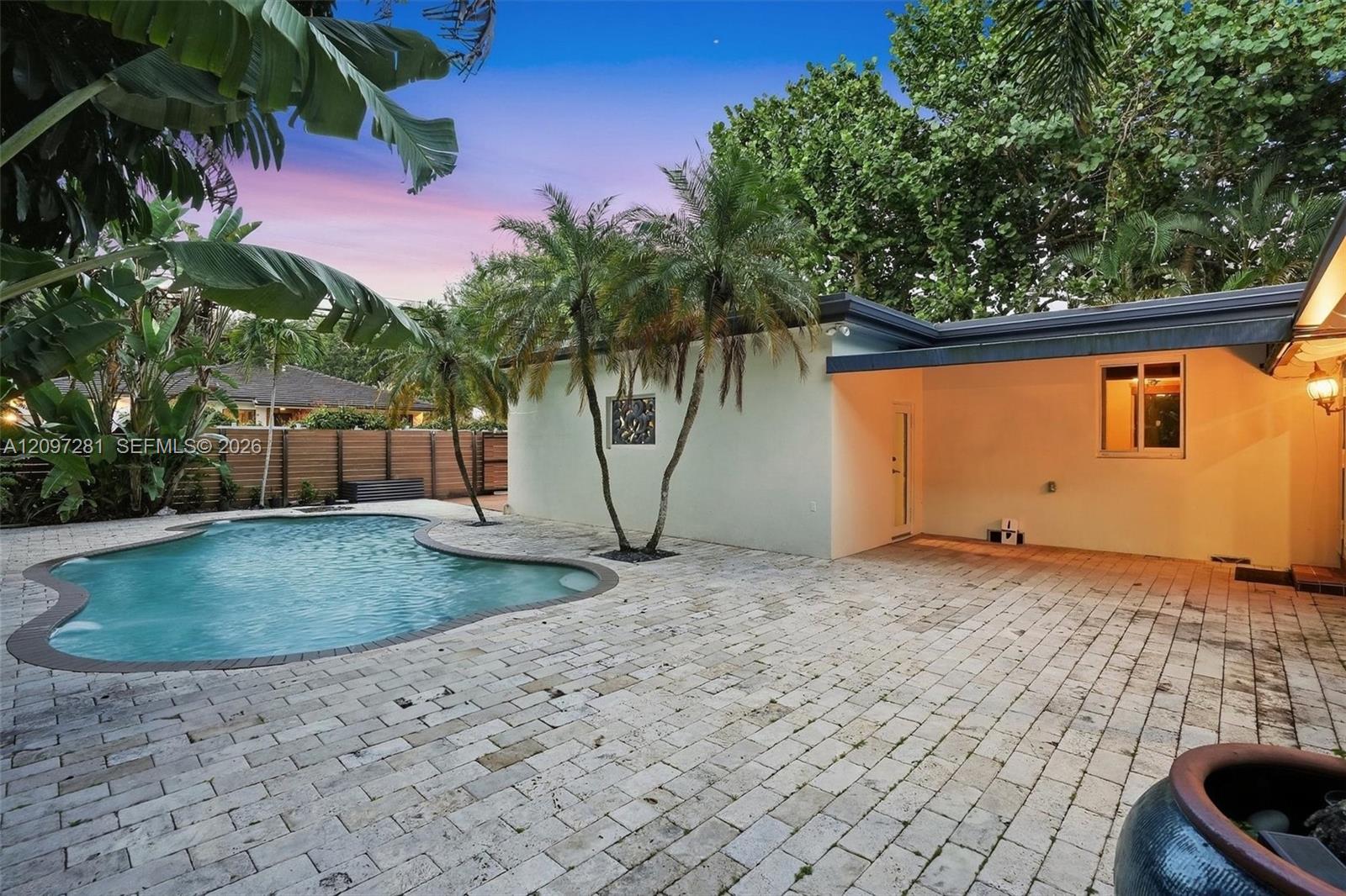 Back patio with a curved swimming pool, pale pavers, and tropical landscaping at dusk.