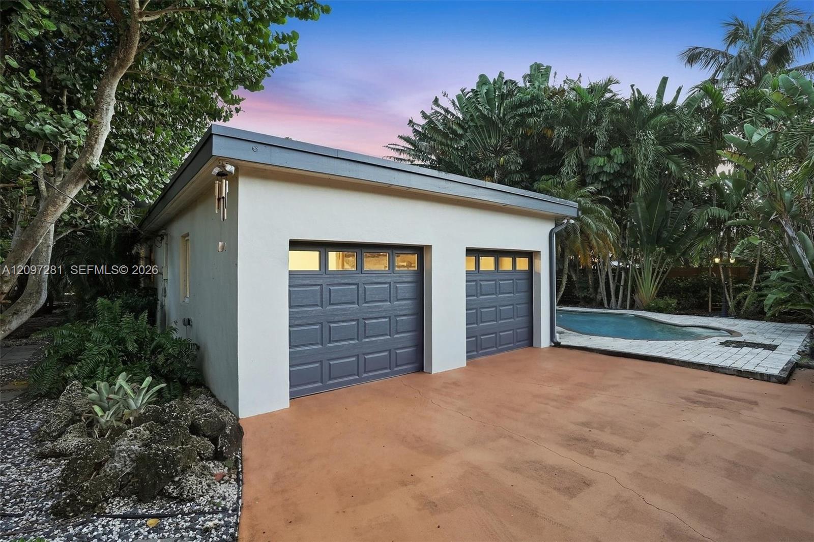 Detached two-car garage with gray doors beside a pool and tropical landscaping.