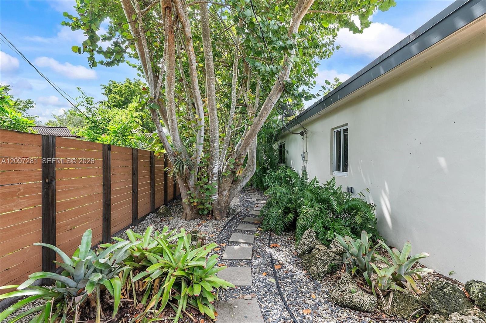 Side yard with a gravel path, tropical plants, wooden fence, and white house exterior.