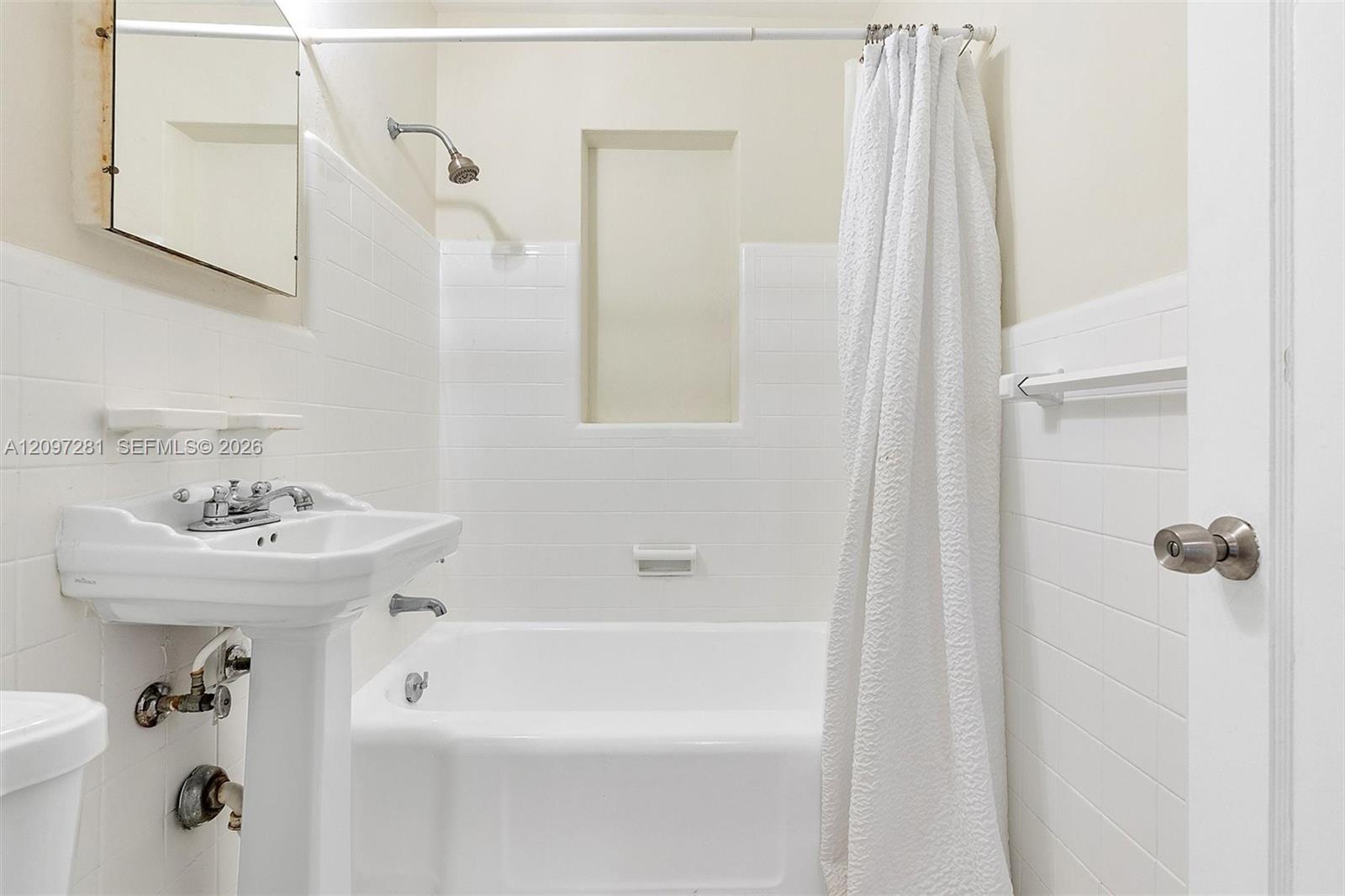 White-tiled bathroom with a bathtub, shower curtain, pedestal sink, and mirrored medicine cabinet.