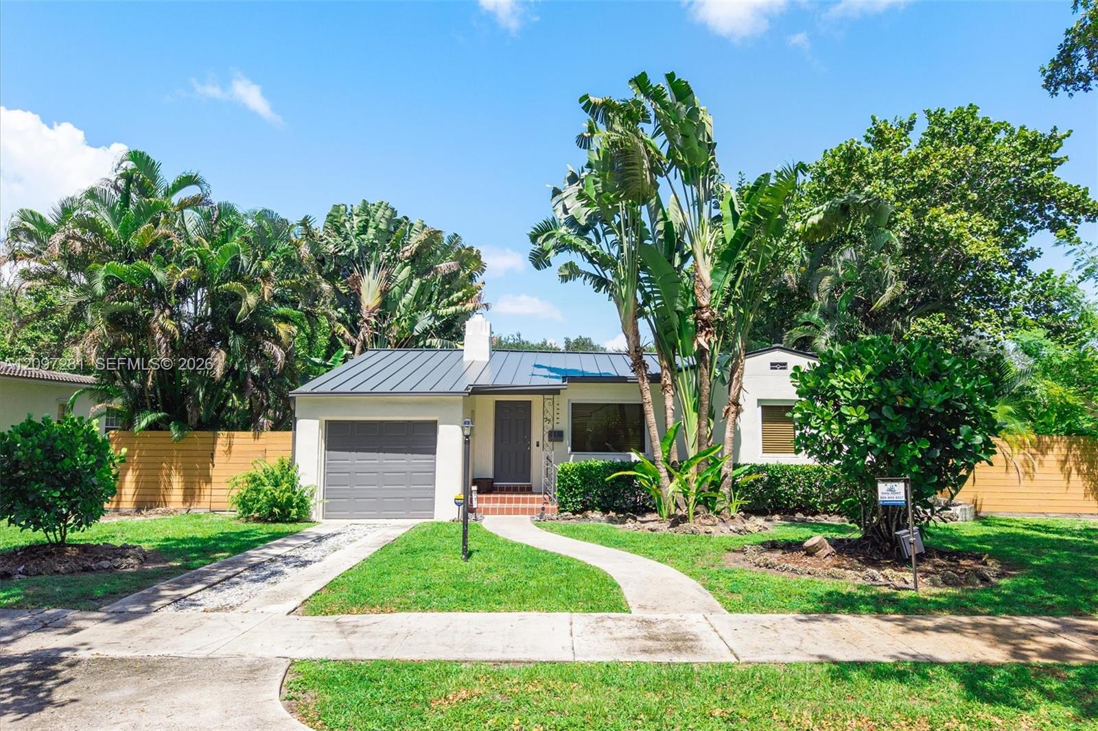 Single-story home with a gray metal roof, garage, and tropical landscaping.