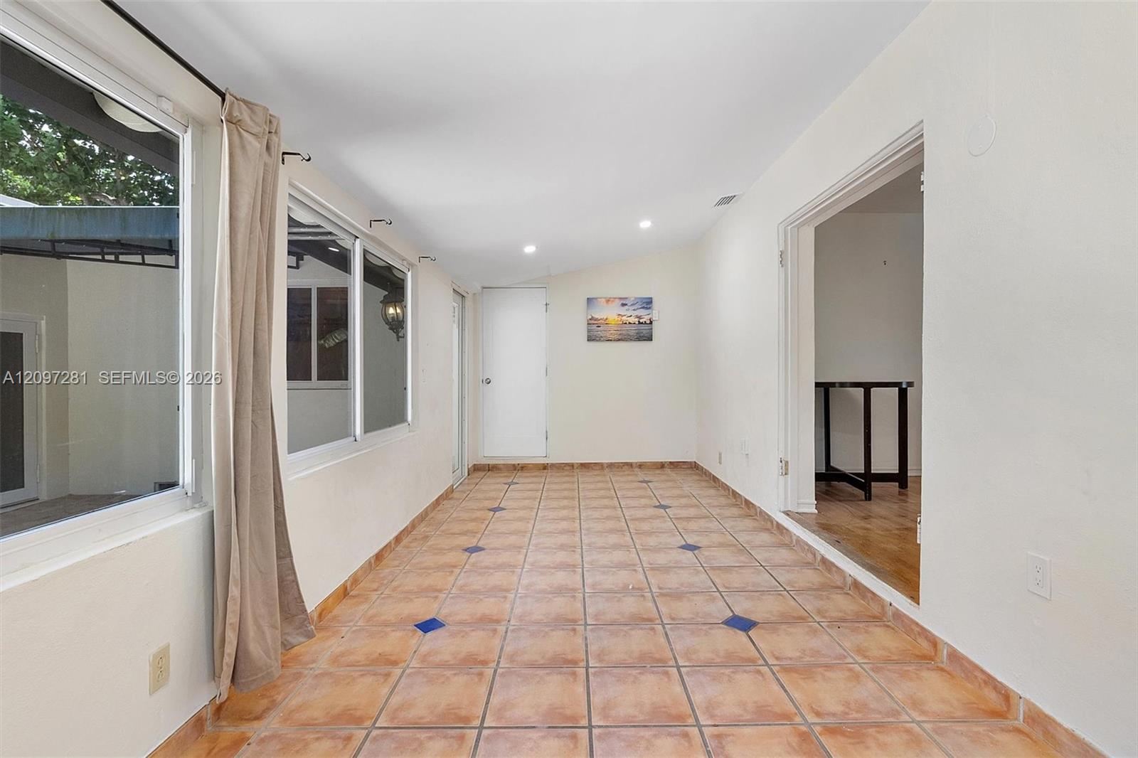 Empty sunroom with terracotta tile floors, large windows, and beige curtains.