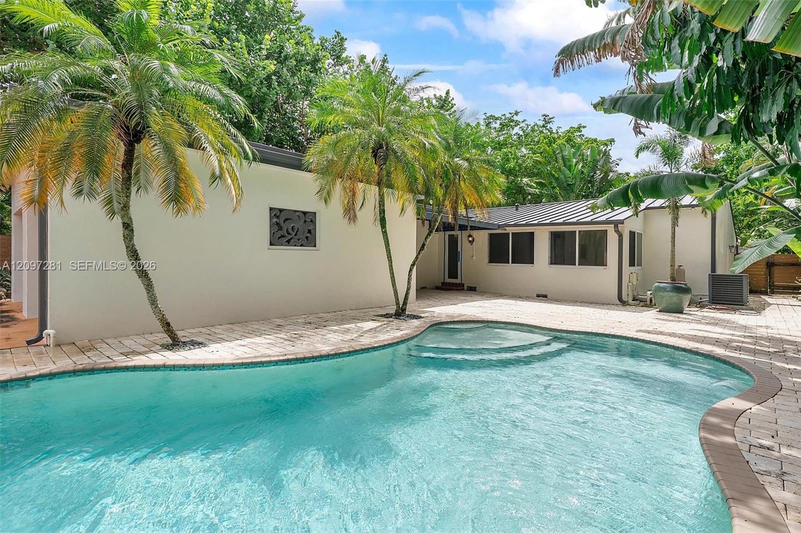 Backyard pool beside a low white house, surrounded by palms and tropical foliage.