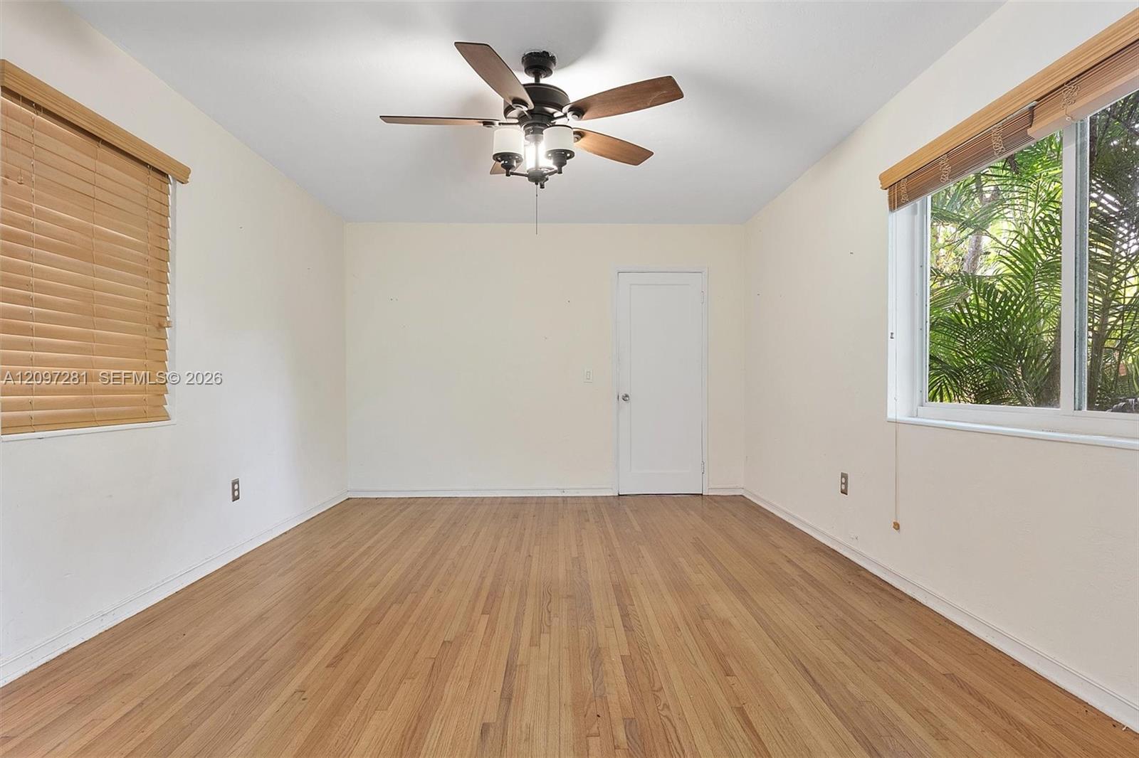Unfurnished bedroom with hardwood floors, a ceiling fan, and windows overlooking palm foliage.
