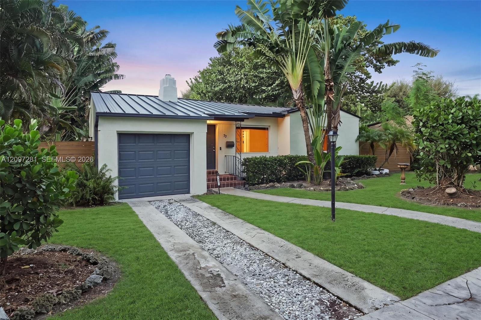 Single-story home with a dark metal roof, blue-gray garage door, and tropical landscaping at dusk.
