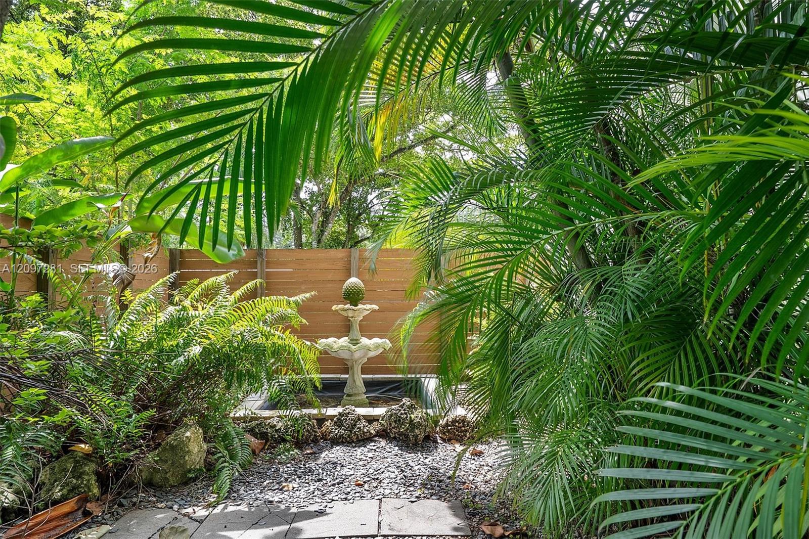 Tropical garden courtyard with a tiered fountain, wooden fence, and gravel path.