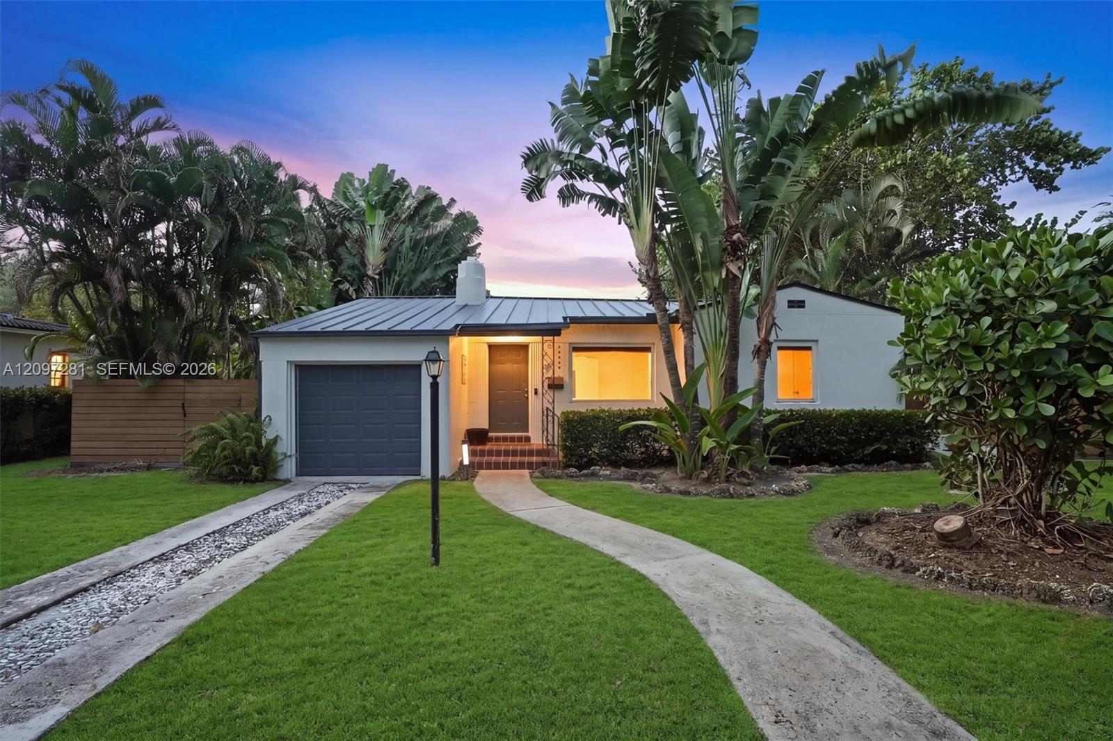 Single-story home with a dark metal roof, attached garage, and tropical landscaping at dusk.