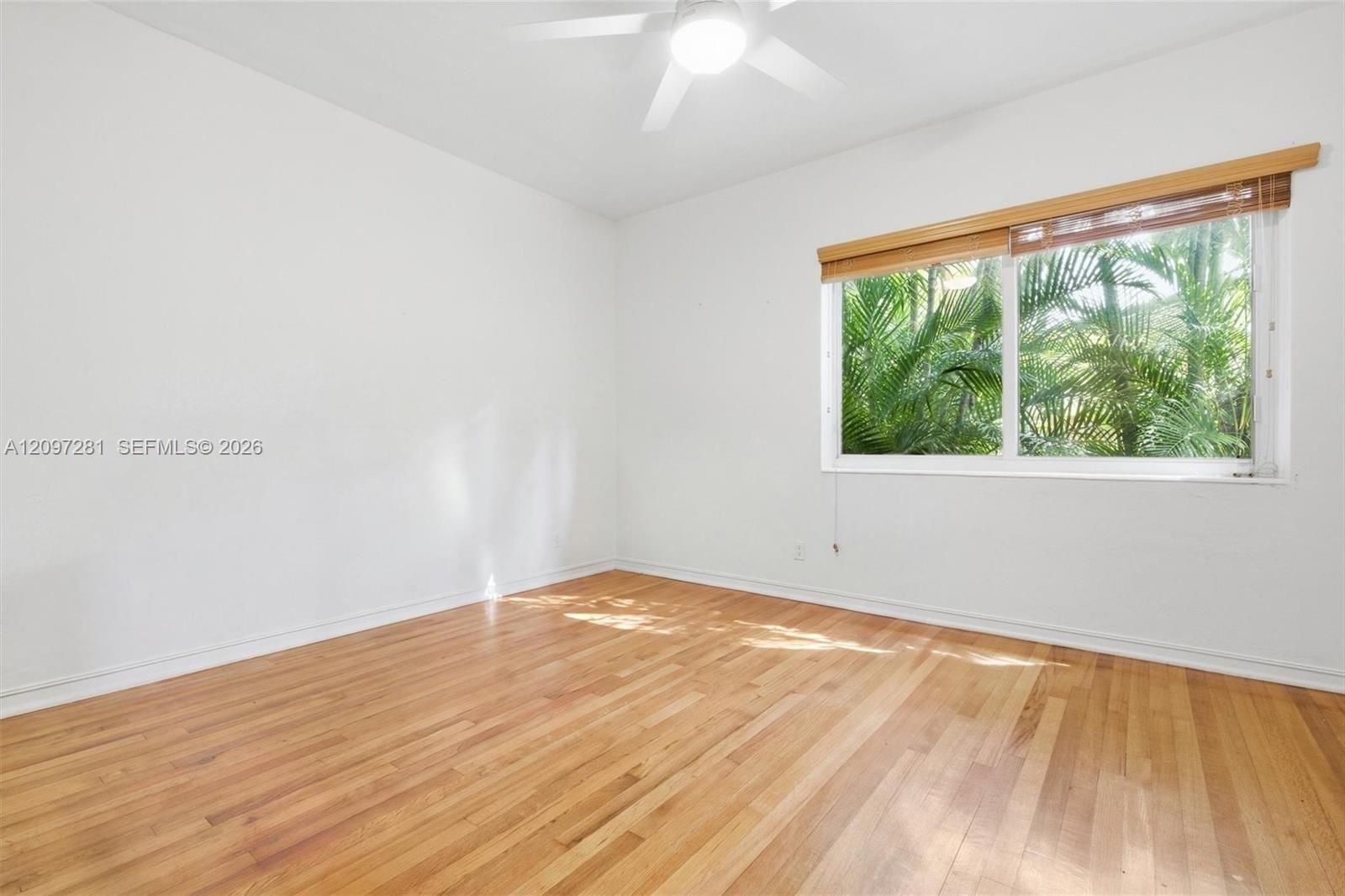 Vacant room with hardwood floors, ceiling fan, and a wide window overlooking tropical foliage.