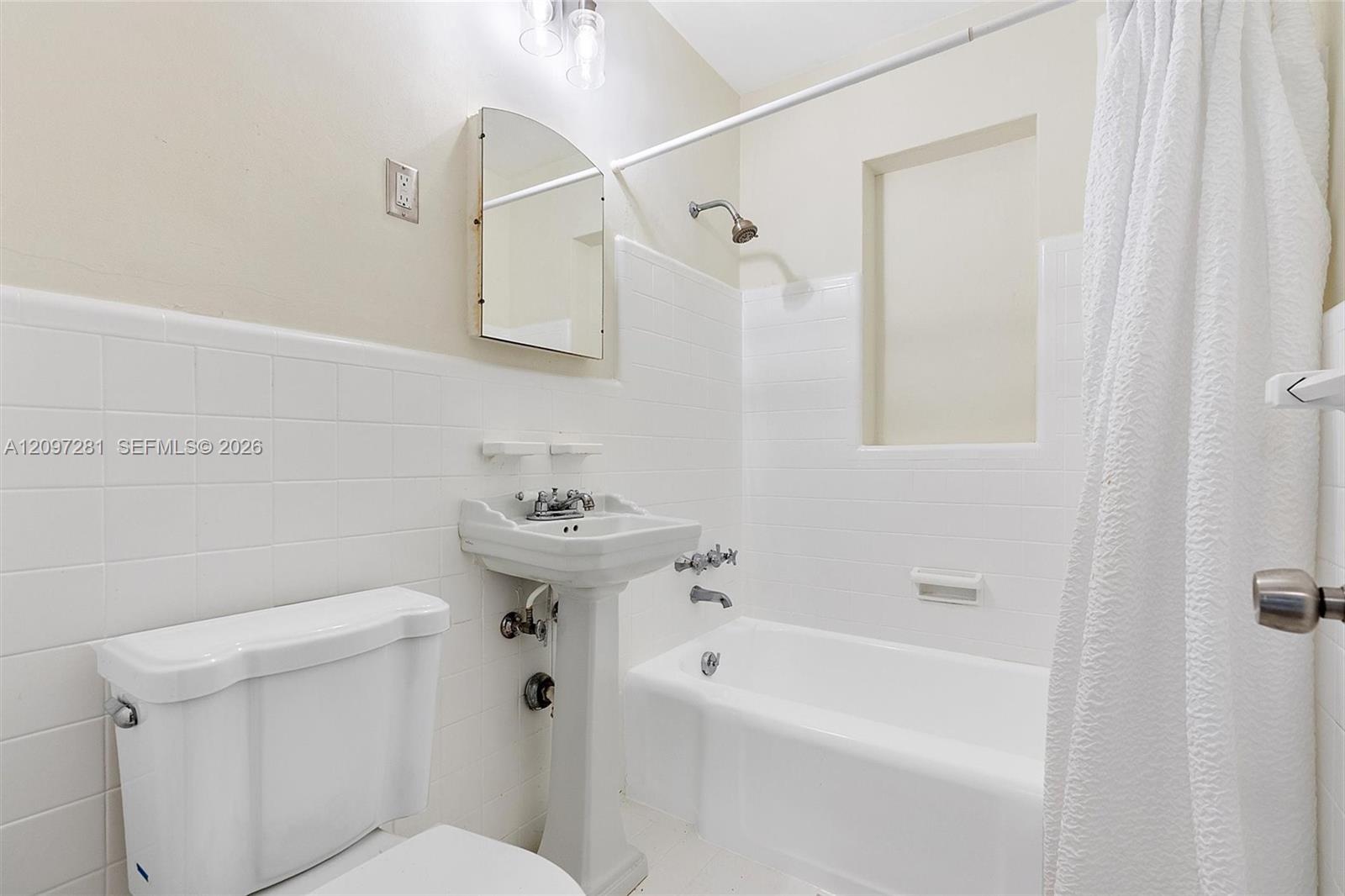 White-tiled bathroom with a bathtub, shower curtain, pedestal sink, and recessed wall niche.