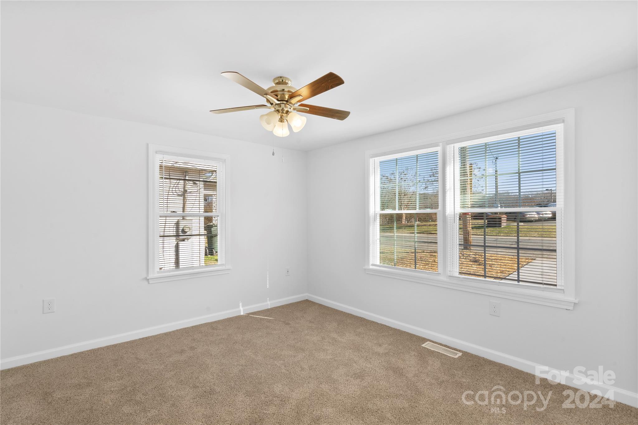 Empty carpeted room with white walls, three blind-covered windows, and a brass ceiling fan.