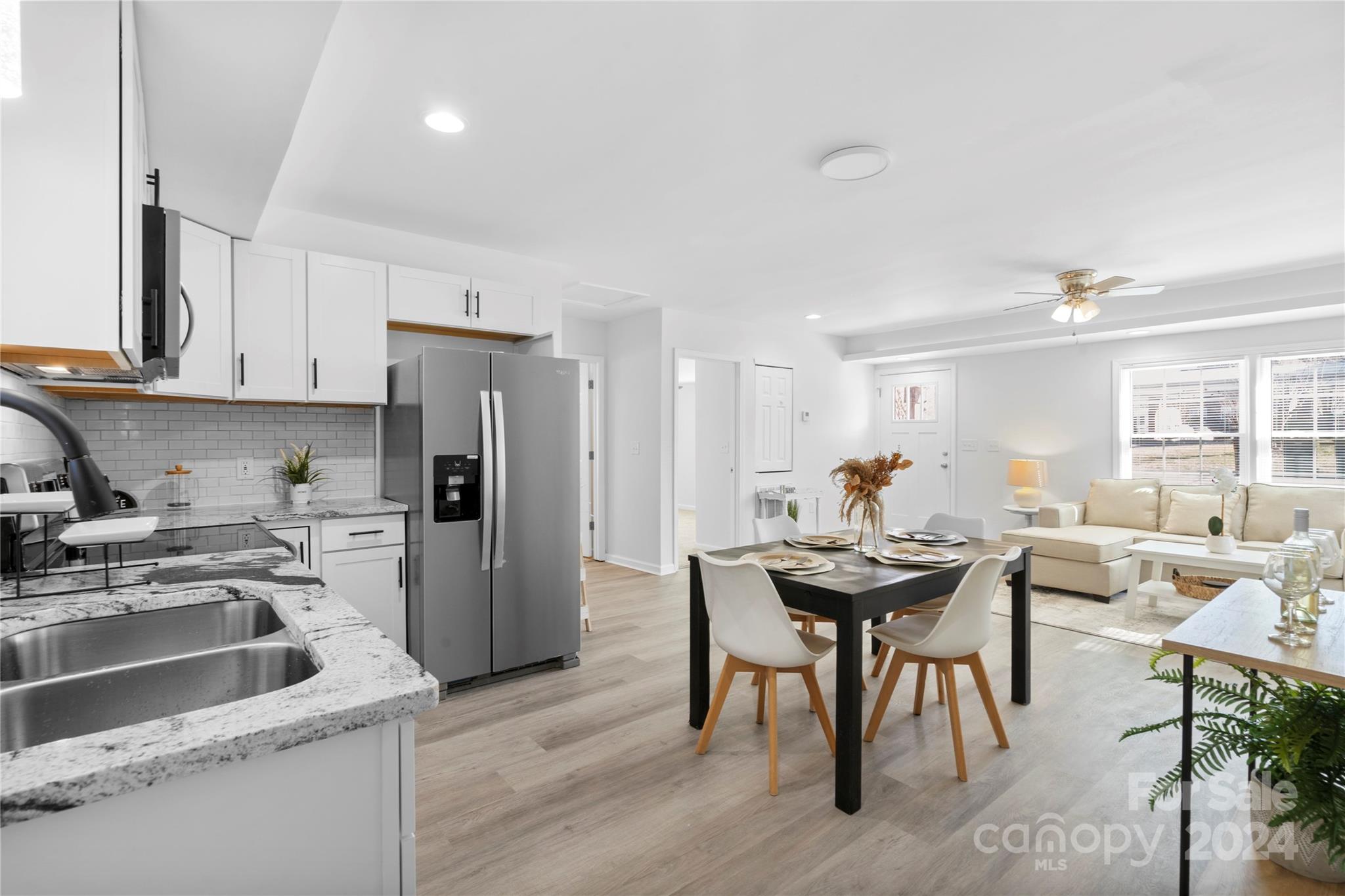 Open kitchen and living area with white cabinets, granite counters, and a dining table beside a cream sofa.