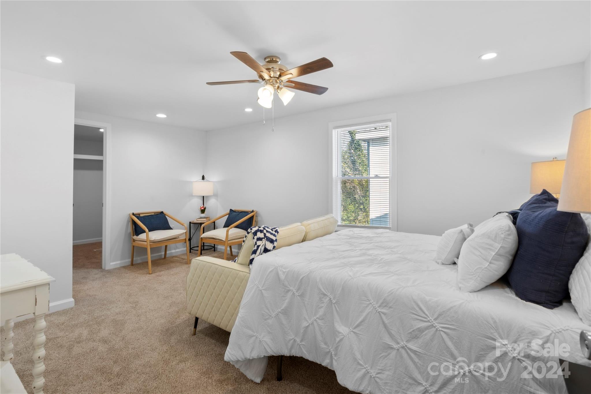 Staged bedroom with a large white bed, ceiling fan, and two armchairs beside a window.