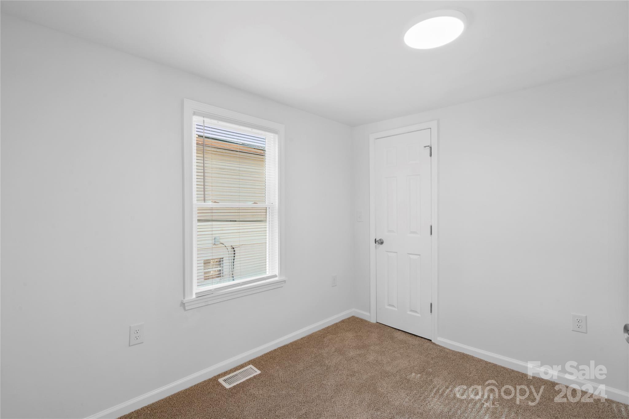 Empty carpeted room with white walls, a window with blinds, and a closed paneled door.