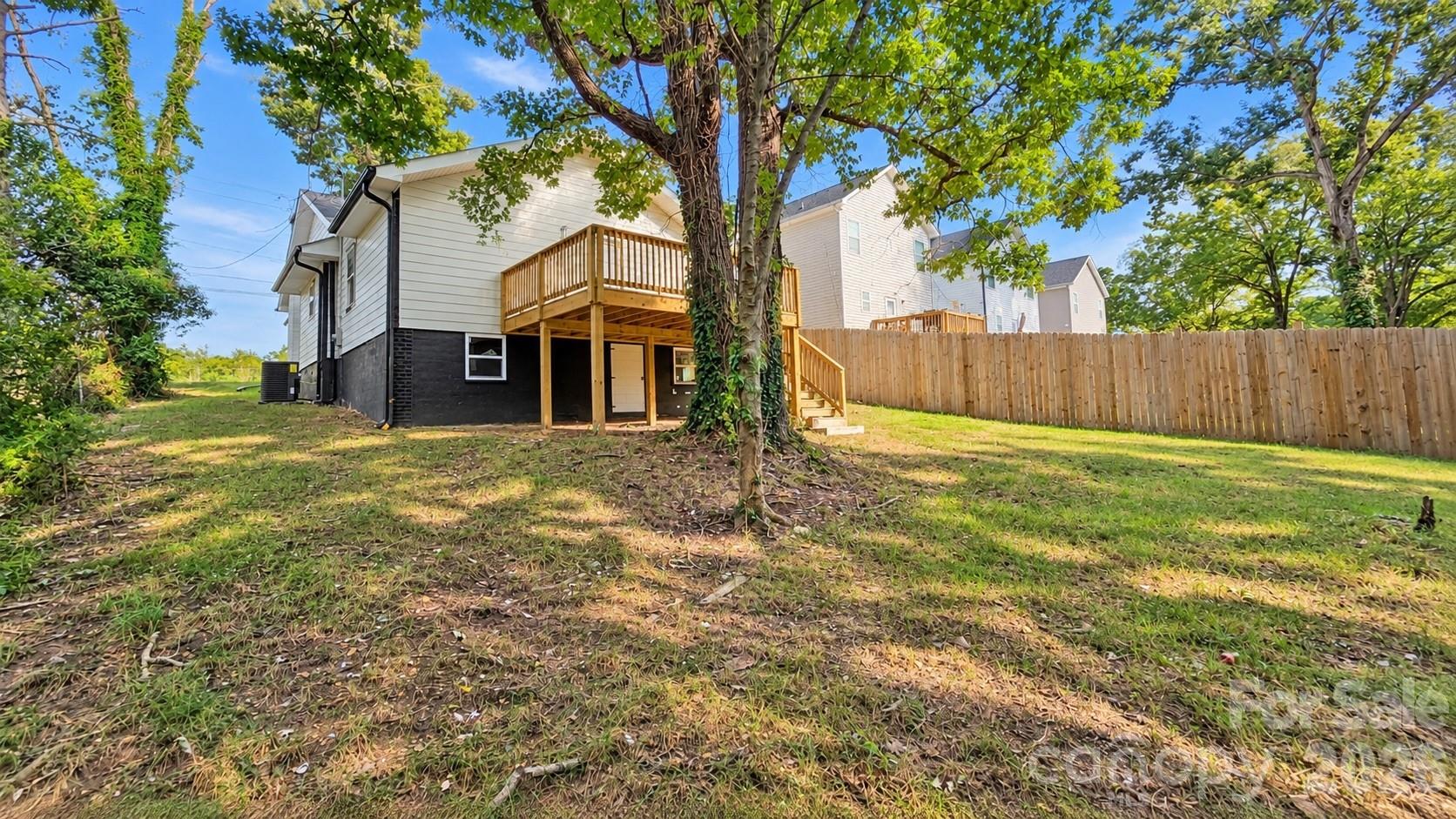 Rear of a light-sided house with a raised wooden deck and fenced, tree-shaded backyard.
