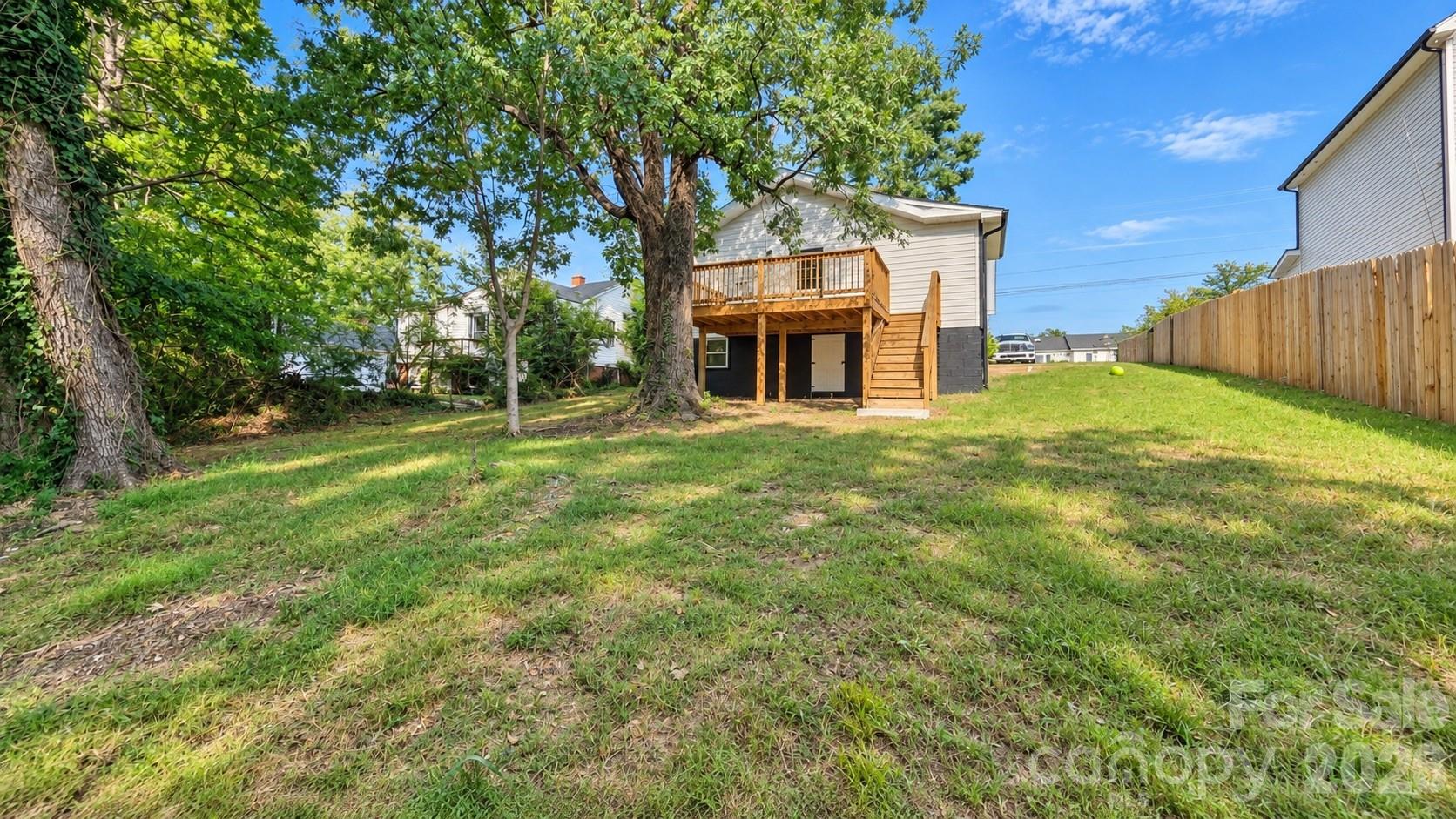 House rear with raised wooden deck and stairs in a tree-shaded grassy backyard.