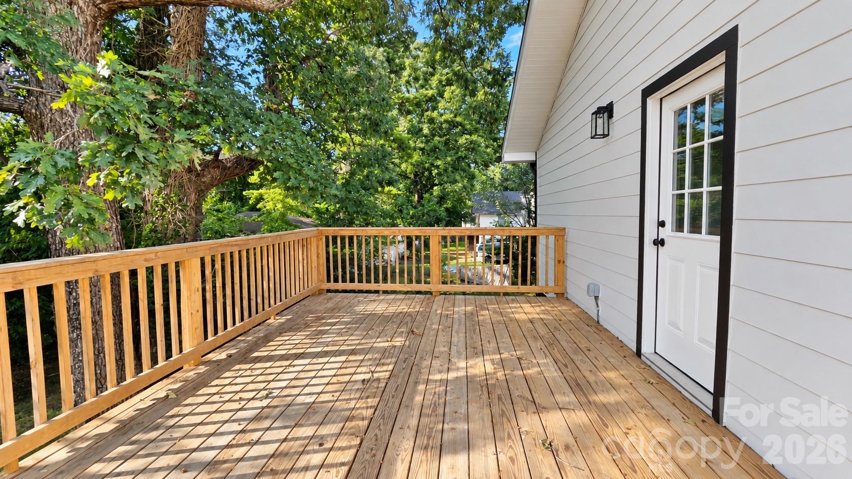 Spacious wooden deck with railings beside a white-sided home, shaded by mature trees.