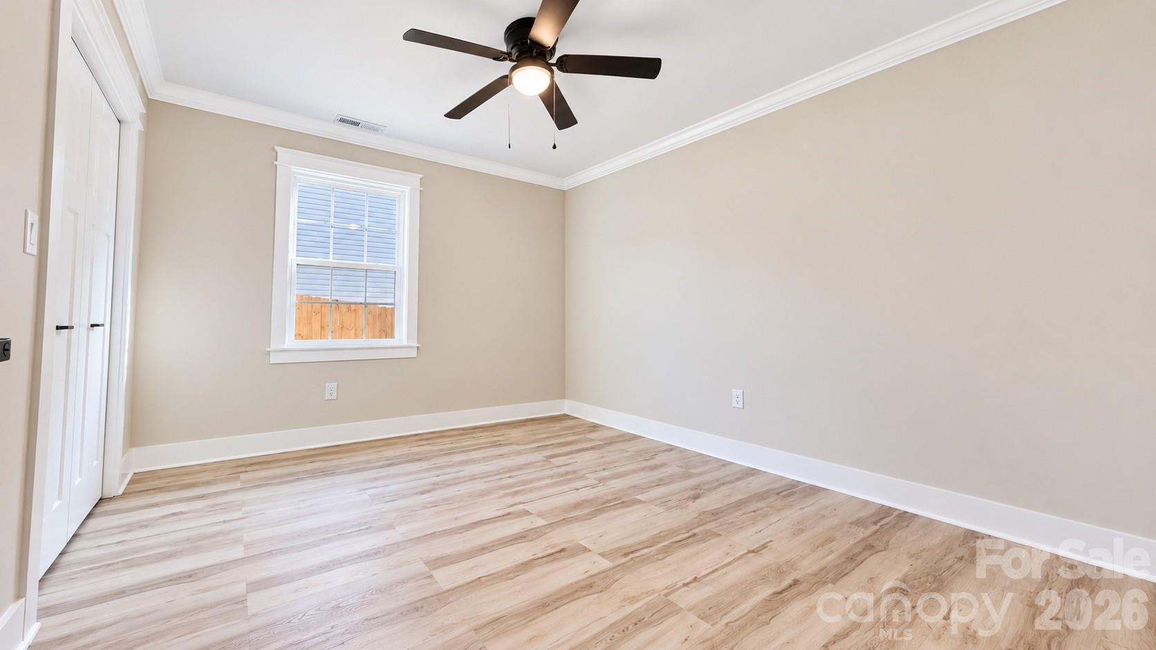 Empty bedroom with light wood-look flooring, beige walls, a window, and a ceiling fan.