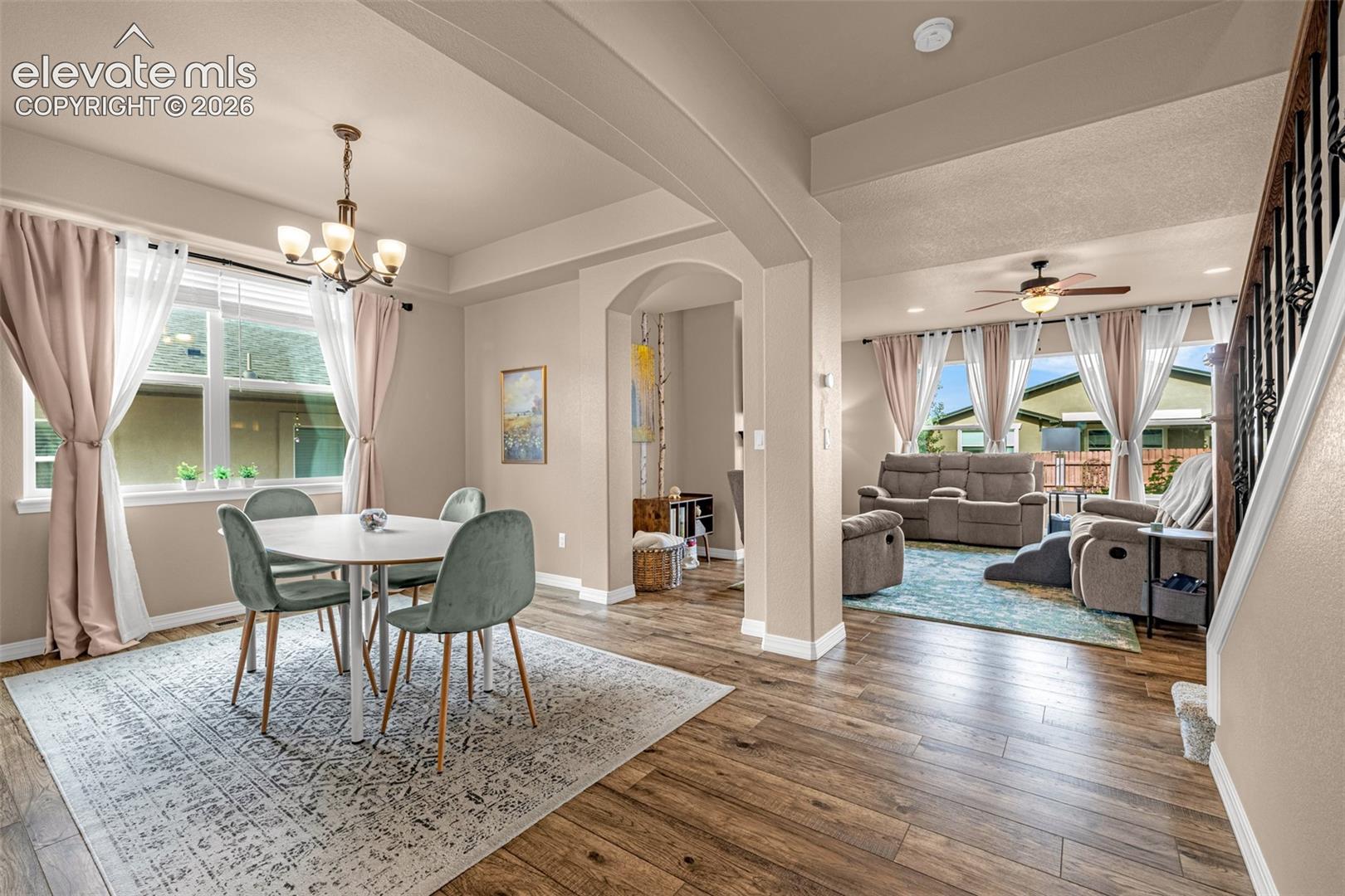 Furnished dining and living rooms connected by an arch, with large curtained windows and wood-look floors.
