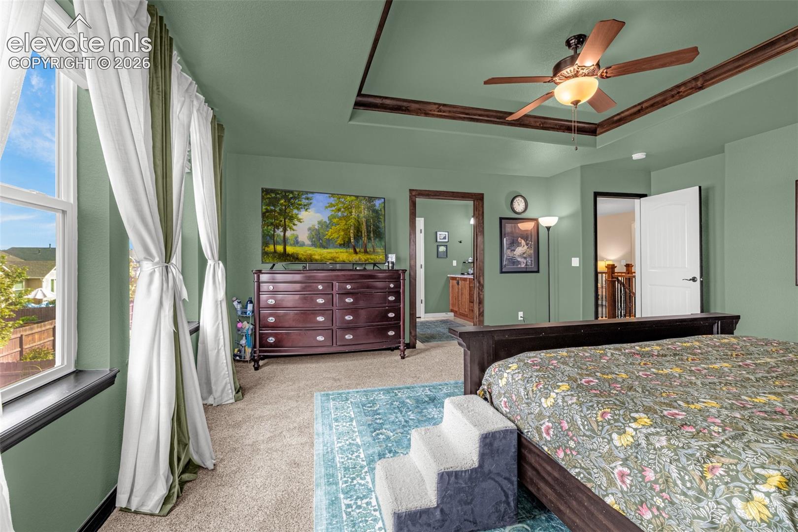 Bedroom with sage-green walls, a tray ceiling, floral bedspread and large curtained windows.
