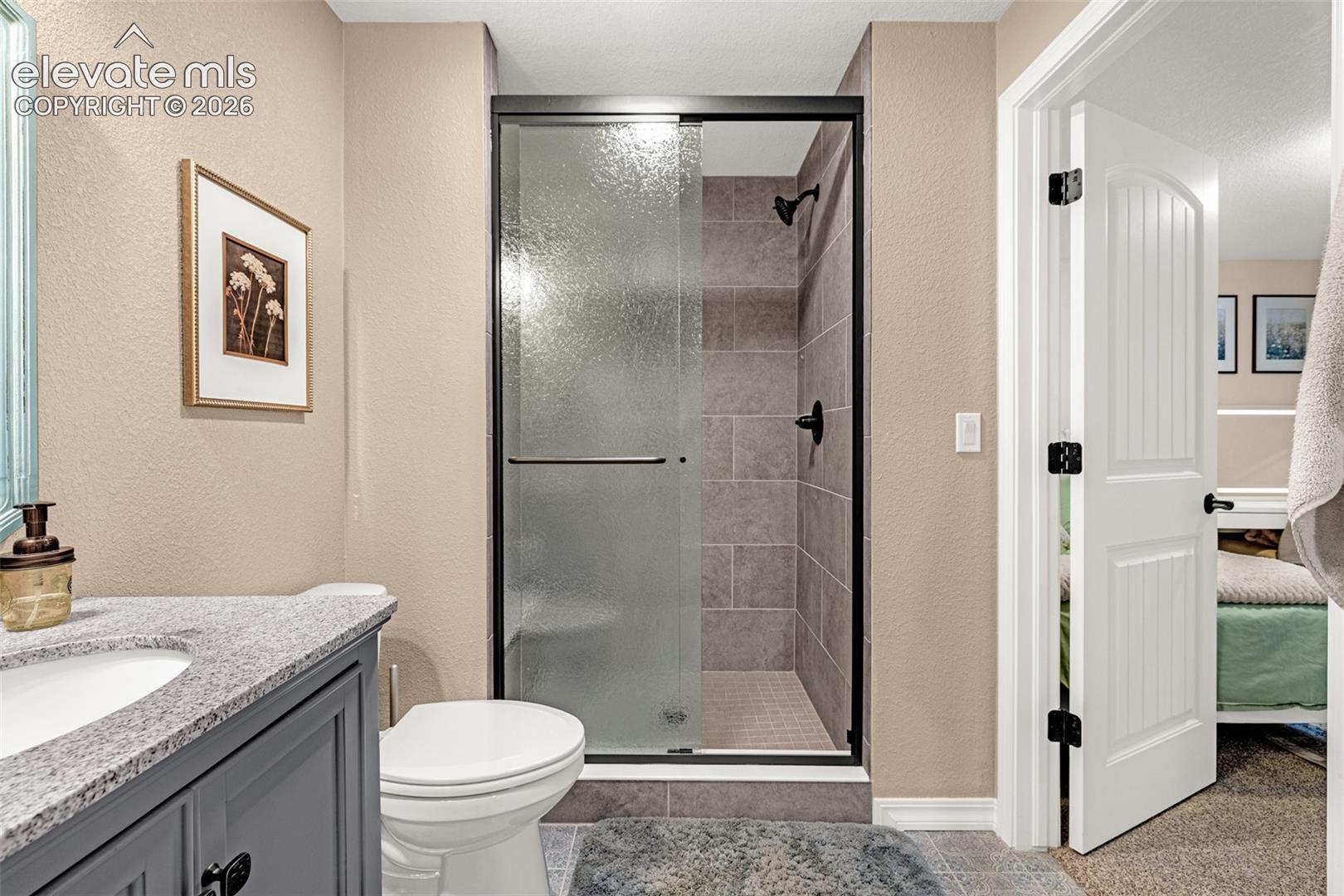 Bathroom with a tiled walk-in shower, gray vanity, and doorway to a bedroom.