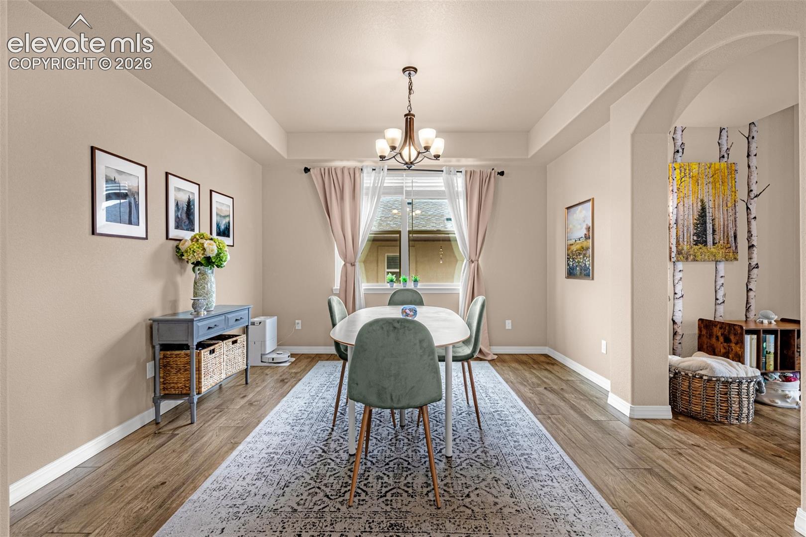 Furnished dining room with an oval table, green chairs, chandelier, and large curtained window.