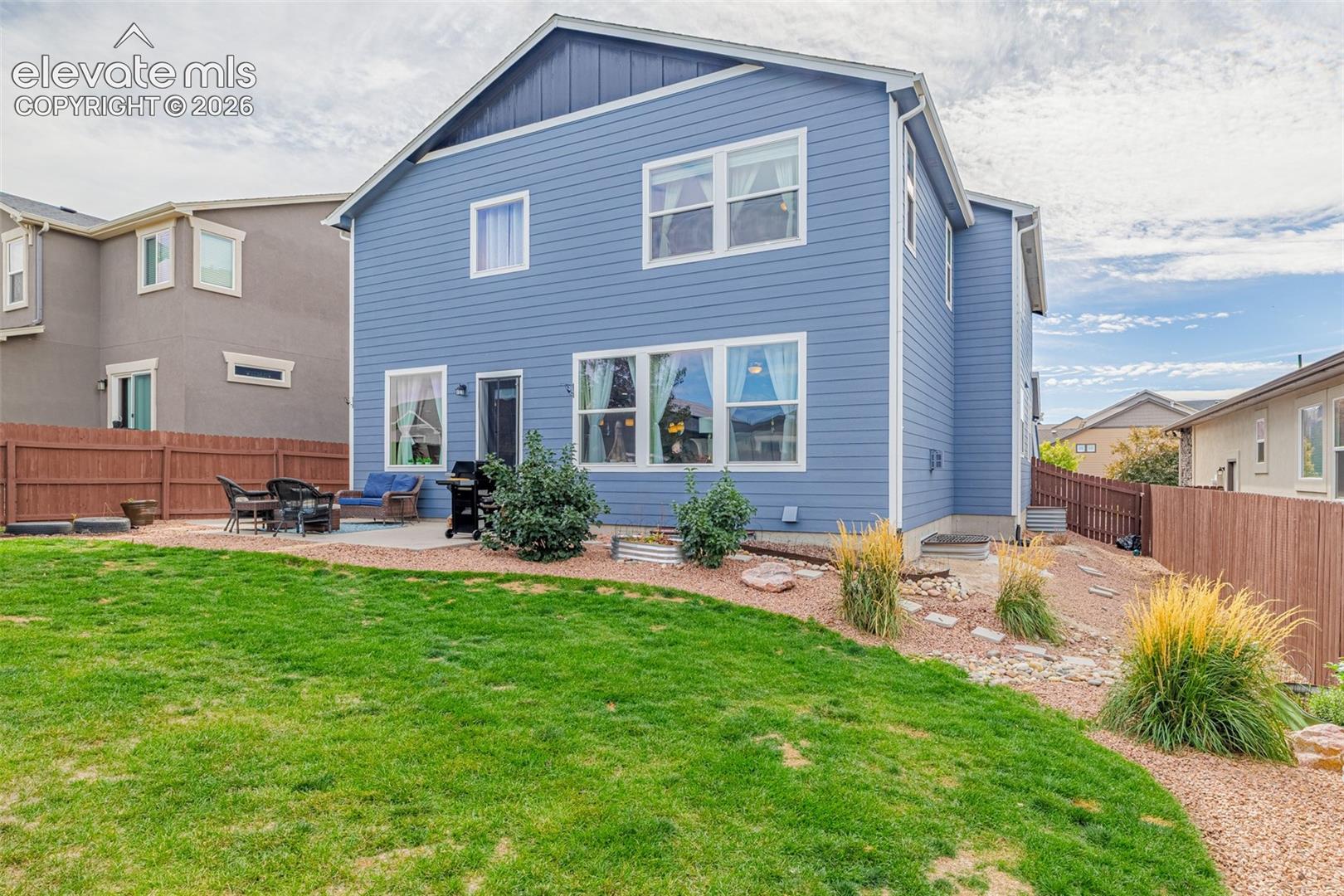 Blue two-story home with a fenced lawn, patio seating, and landscaped gravel beds.