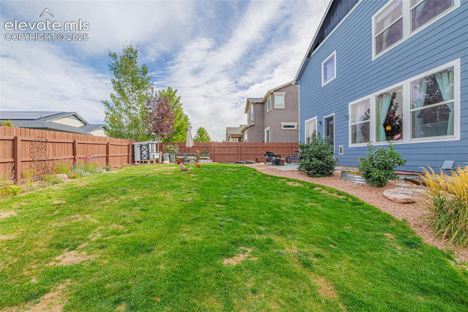 Fenced backyard with a broad lawn, patio seating, and blue-sided house.