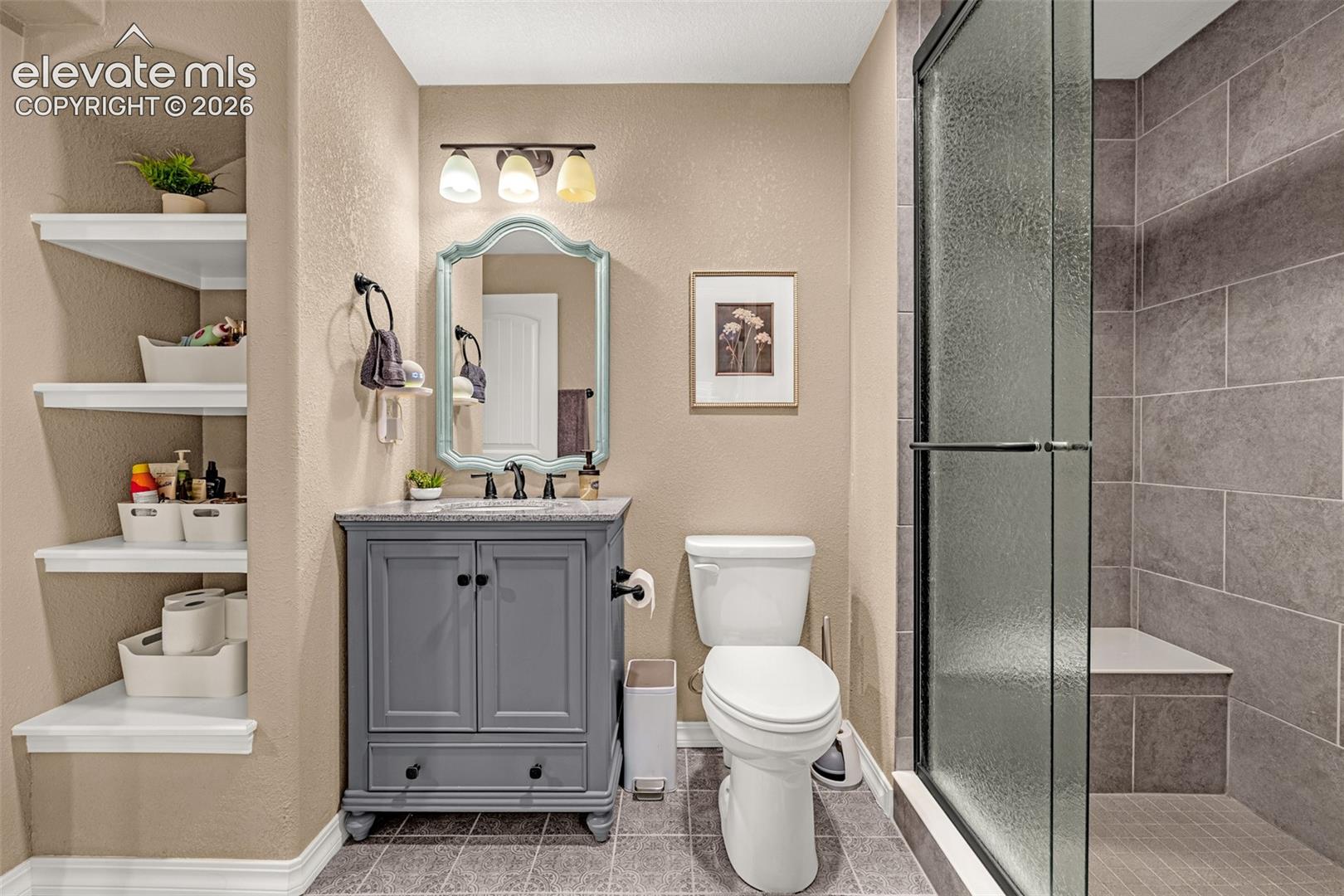 Bathroom with a gray vanity, open storage shelves, toilet, and glass-enclosed tiled shower.