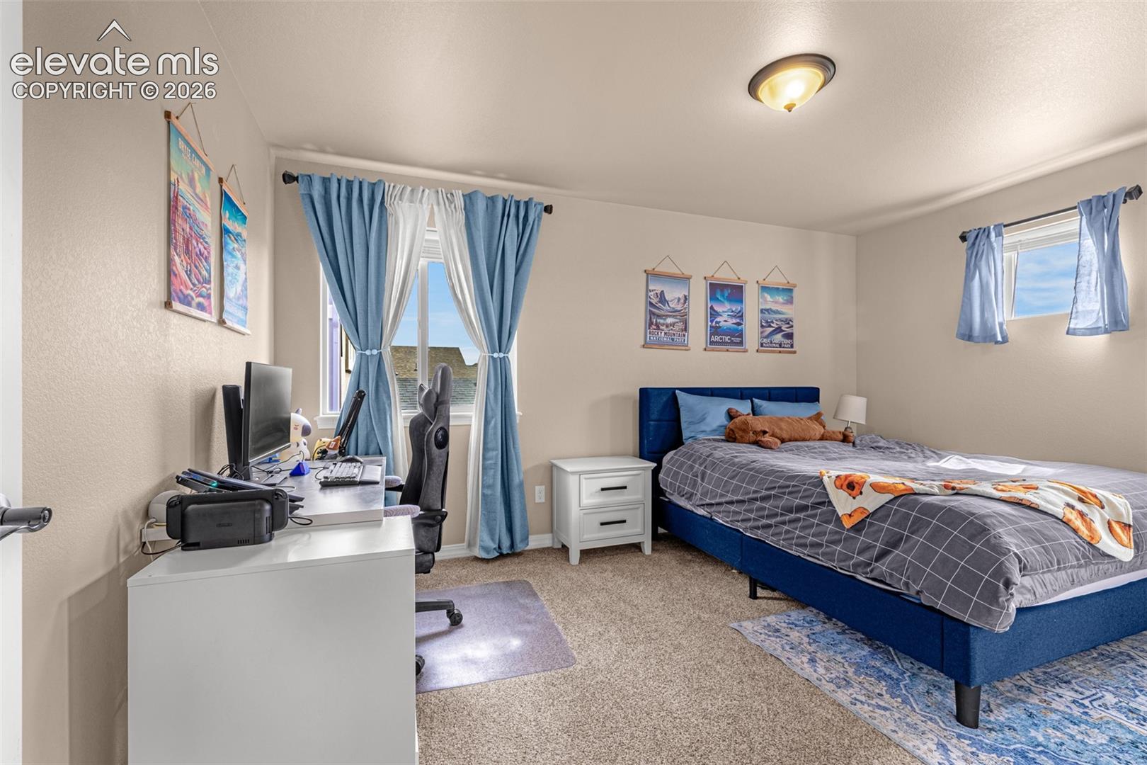 Bedroom with a blue bed, blue curtains, and a computer workstation by the windows.