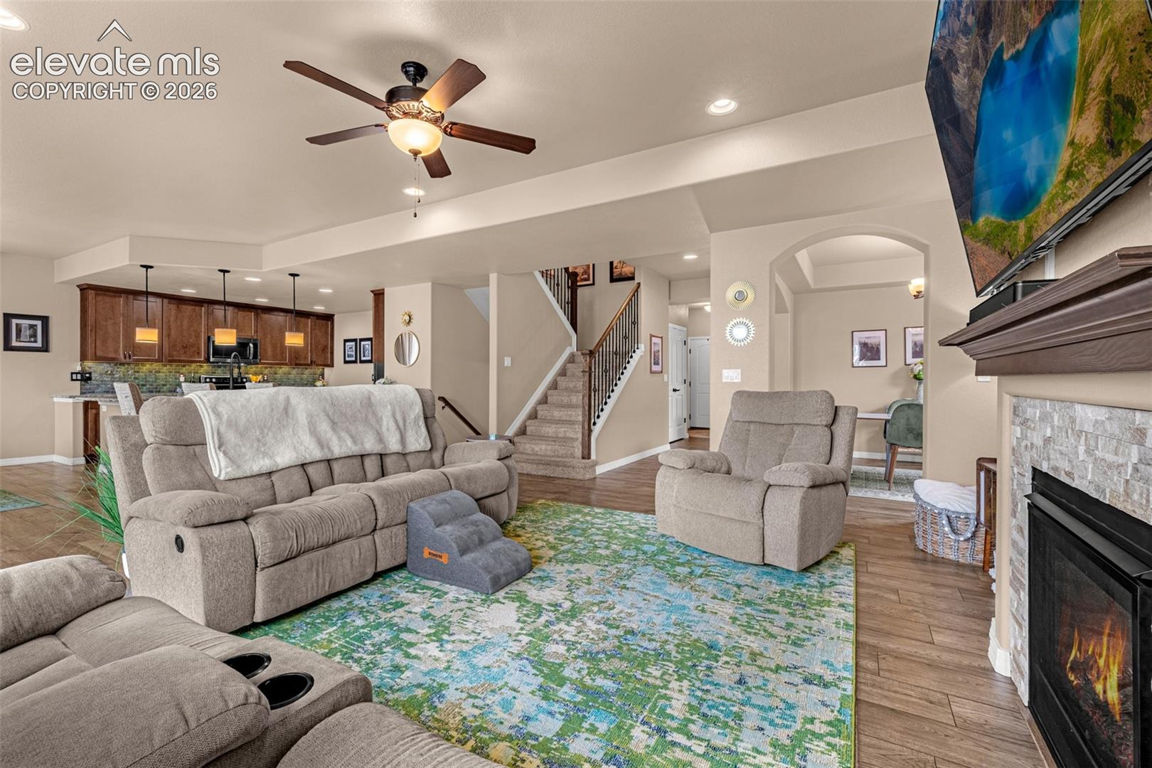 Open-plan living room with reclining sofas, a colorful rug, staircase, and stone fireplace.