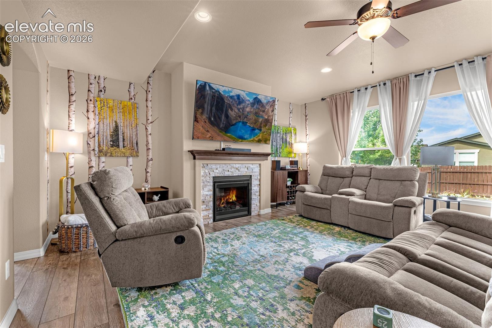 Living room with a stone fireplace, taupe recliners, birch accents, and large curtained windows.