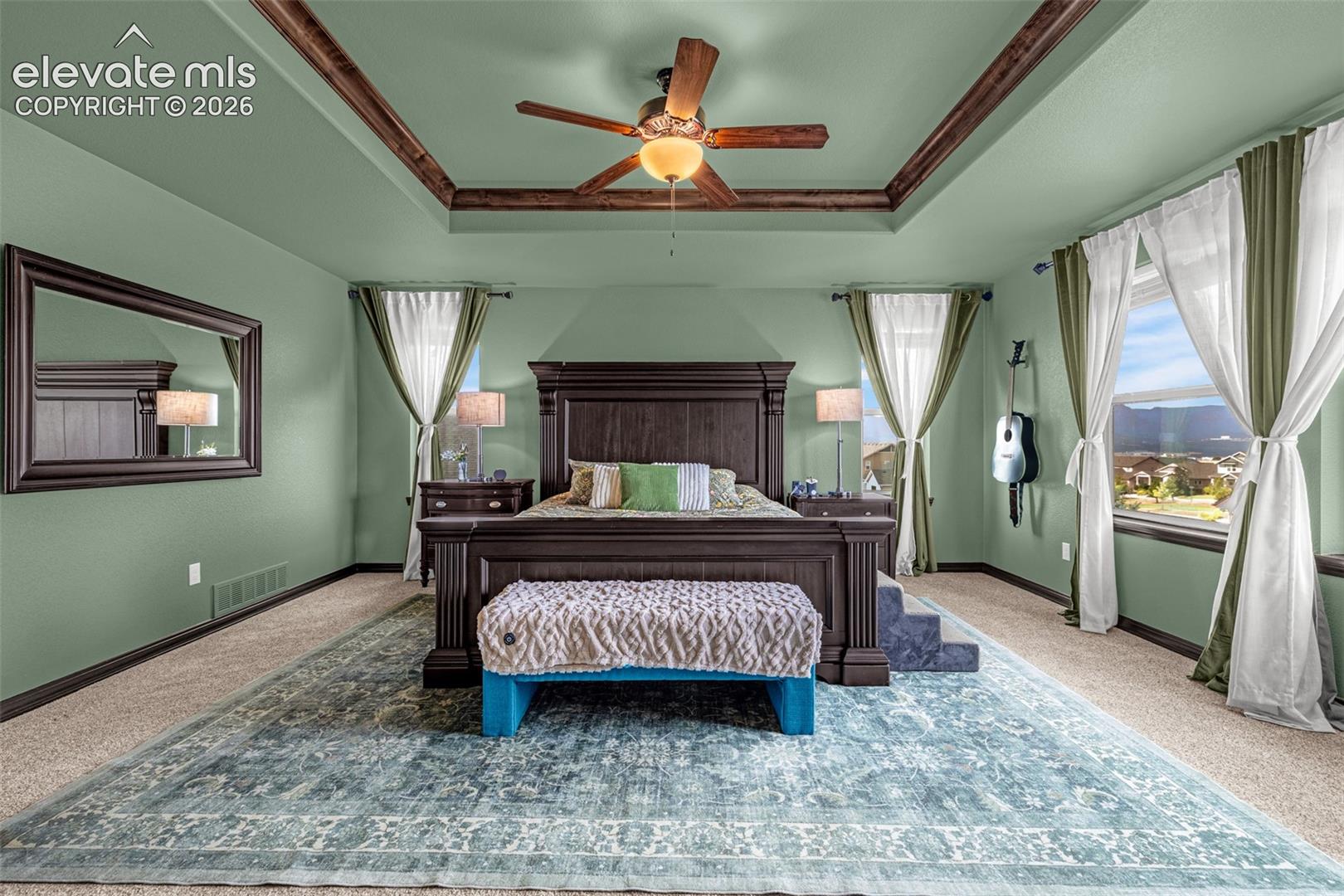 Furnished bedroom with a dark wood bed, tray ceiling, and mountain views through large windows.