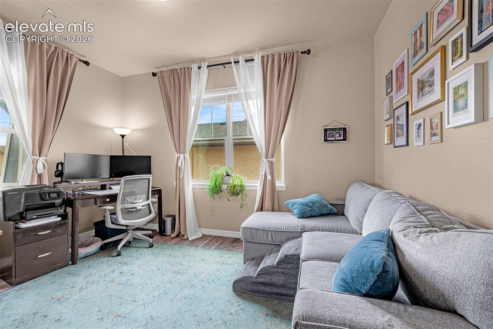 Living room with a gray sectional sofa, blue cushions, and a desk workstation beside curtained windows.