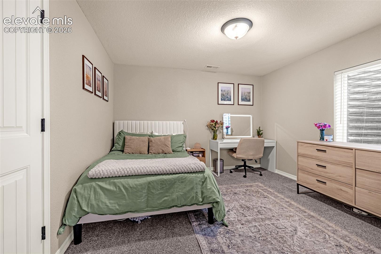 Bedroom with a green-covered bed, desk and chair, and wooden dresser beside a window.