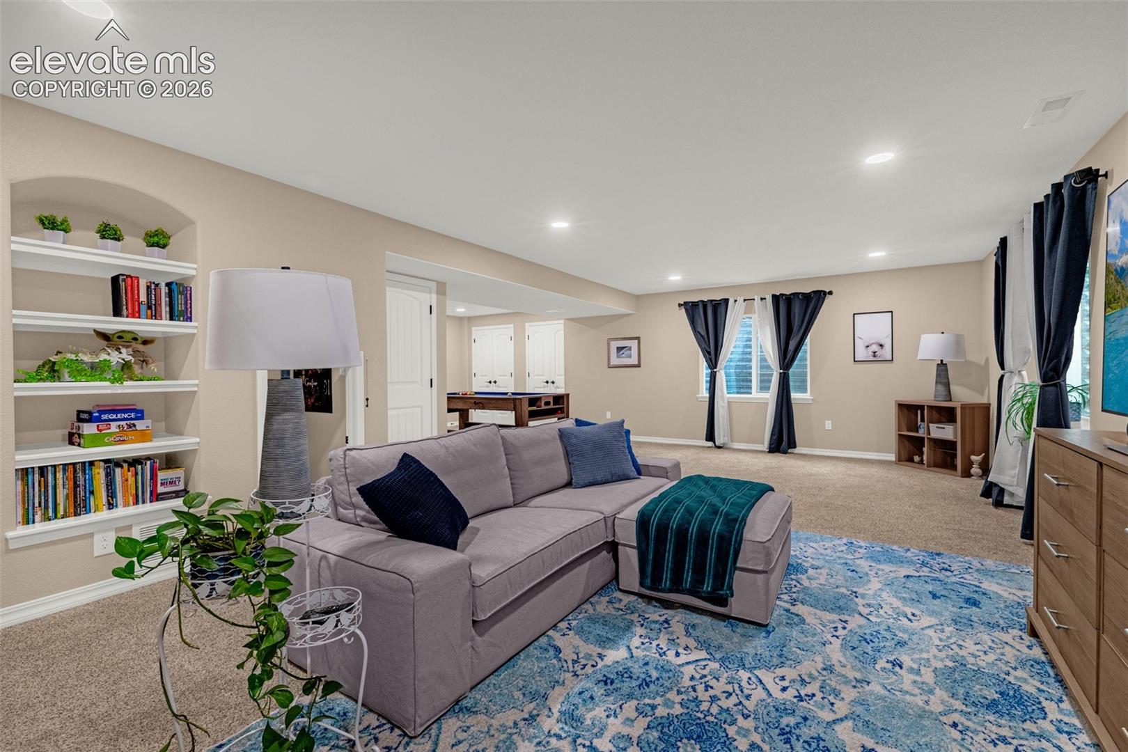 Furnished living room with a gray sectional, blue patterned rug, built-in shelves, and dark curtains.