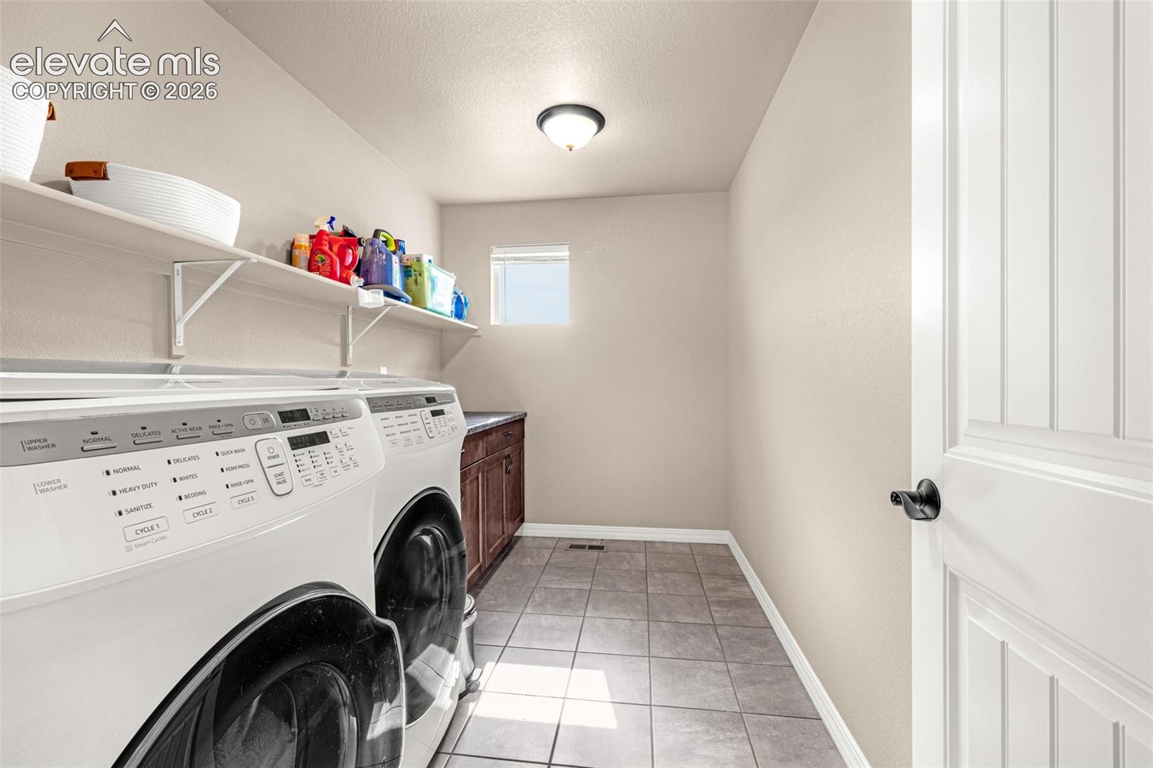 Laundry room with a washer and dryer, supply shelves, and gray tile flooring.
