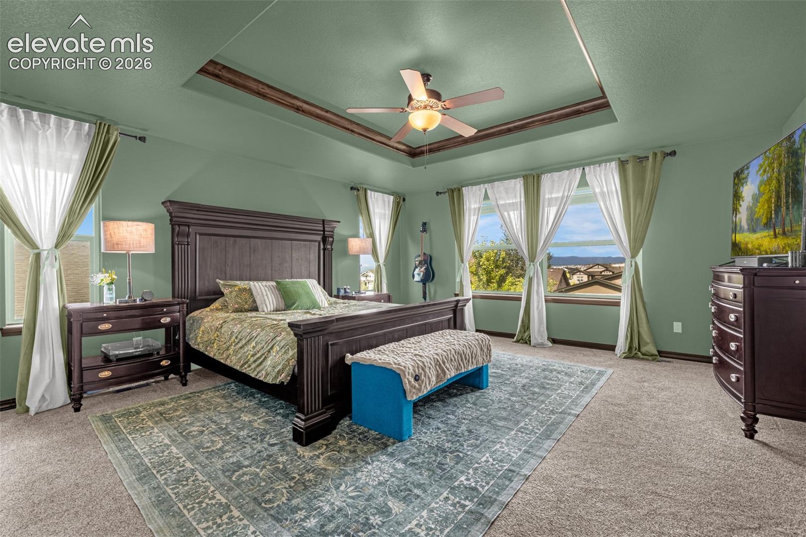 Furnished bedroom with a dark wood bed, tray ceiling, and large windows overlooking distant mountains.