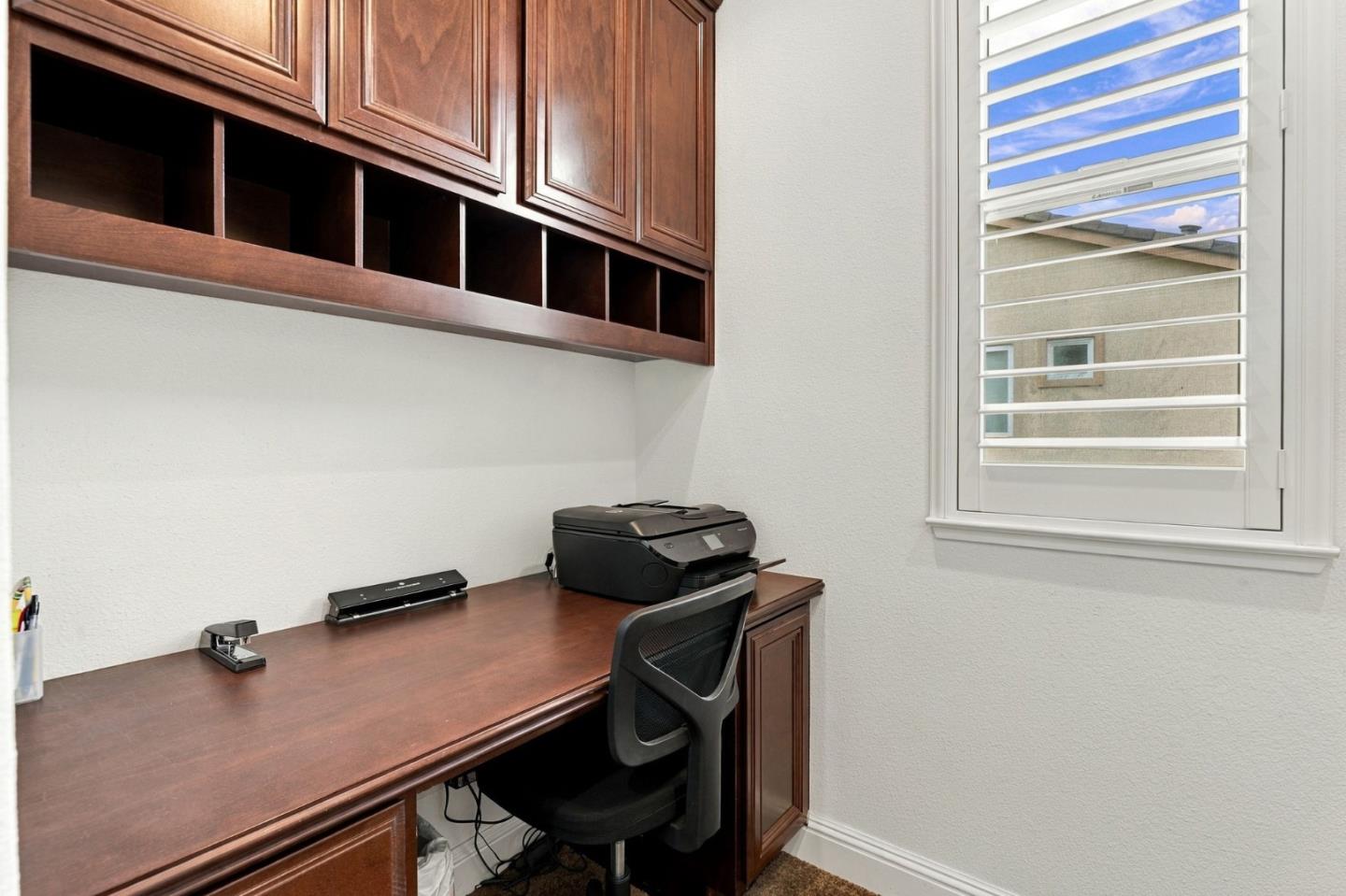 Home office with a built-in dark wood desk and cabinets, black chair, printer, and shuttered window.