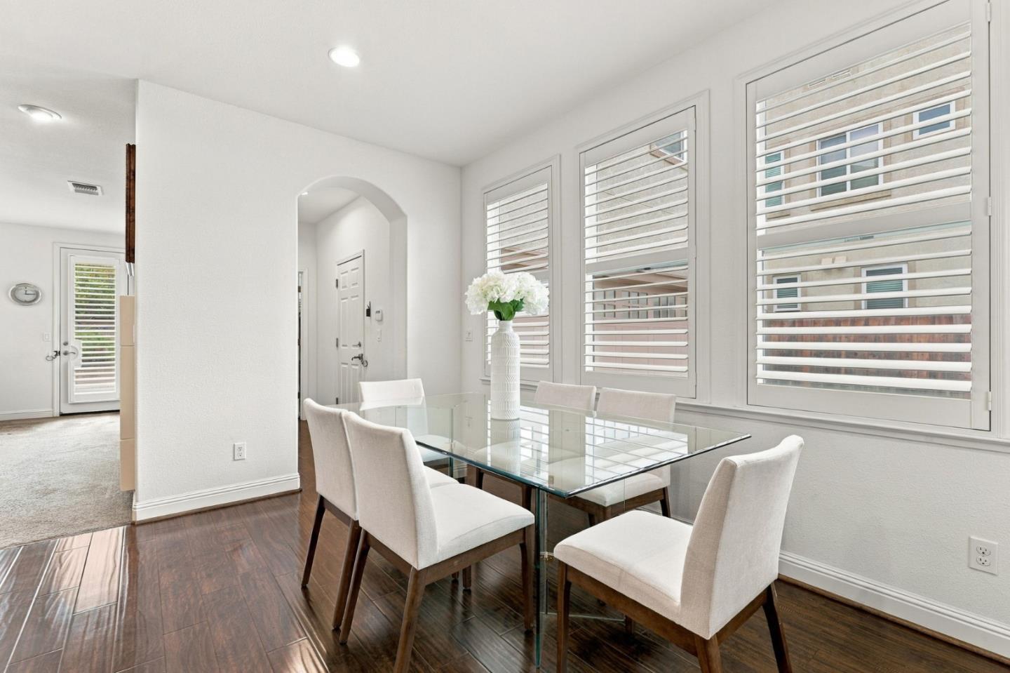 Dining area with a glass table, upholstered chairs, and tall windows with plantation shutters.